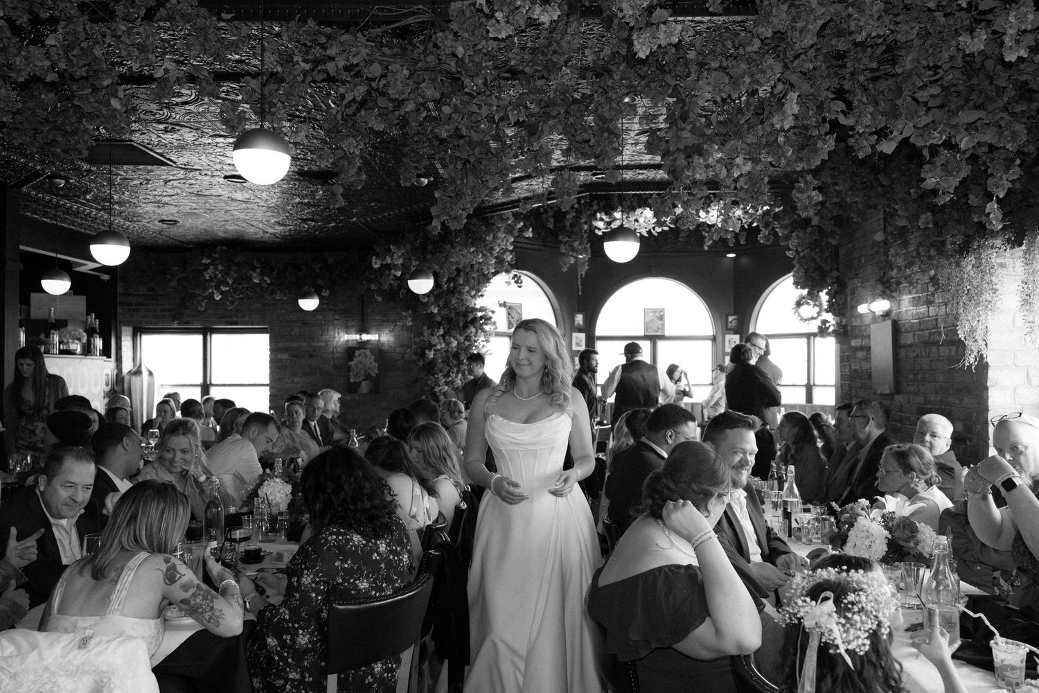 Black and white photo of a bride in a white gown standing among wedding reception guests in a decorated restaurant.
