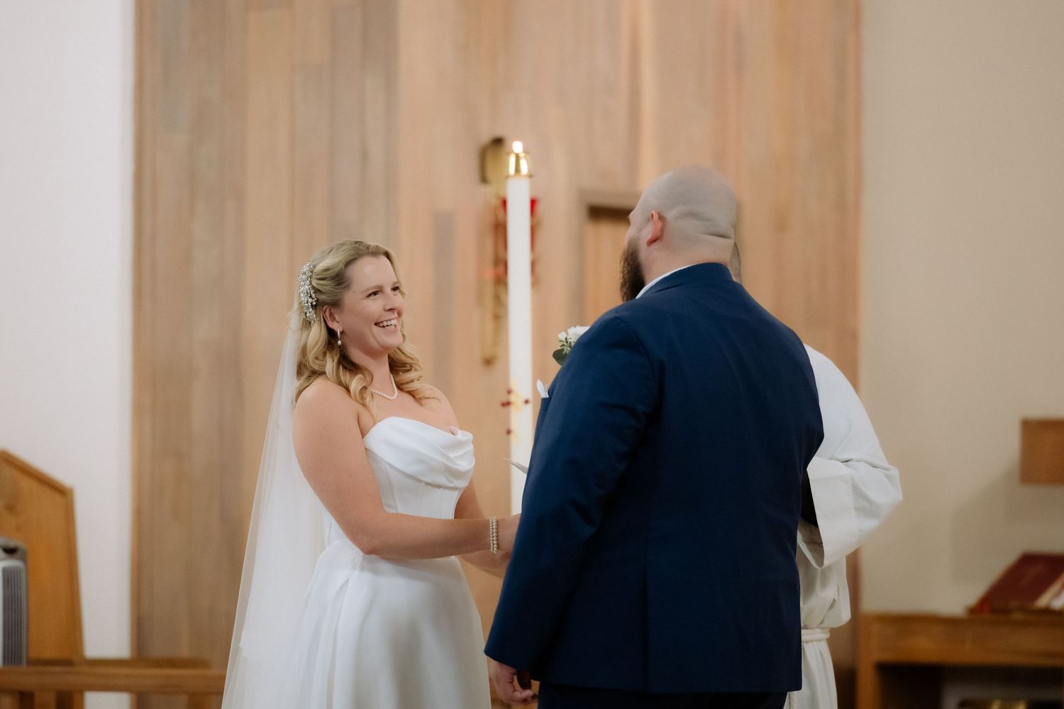 Bride smiling joyfully at groom during wedding ceremony in a church, wearing a white strapless gown and veil.