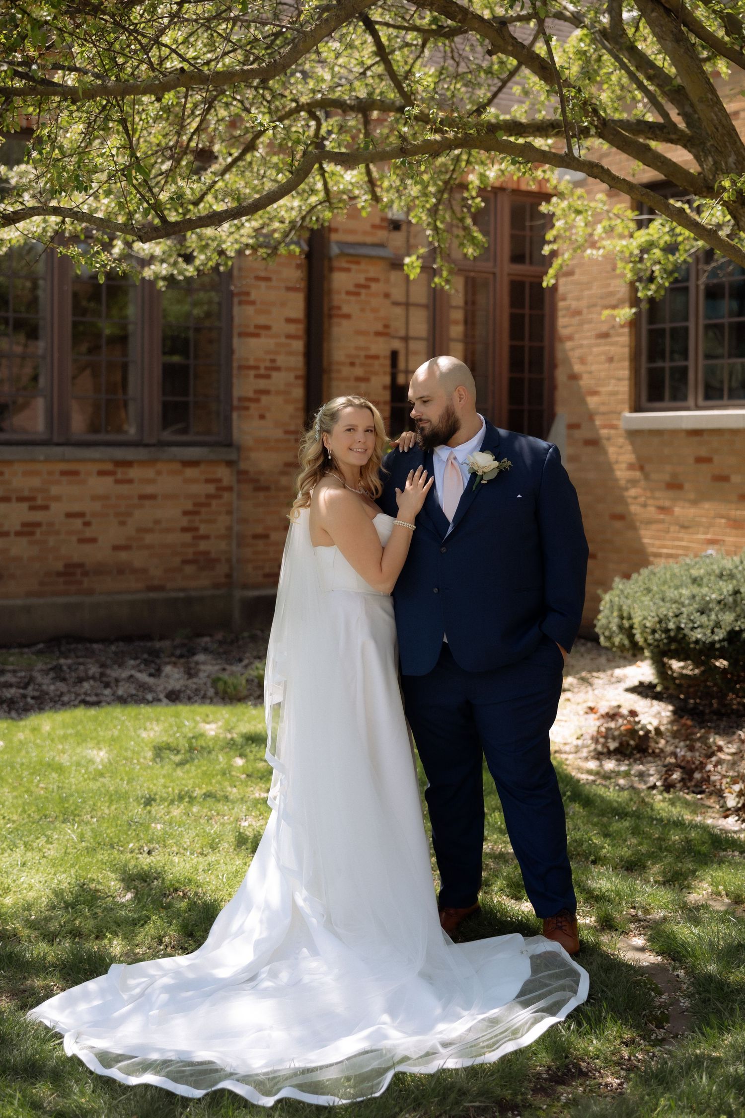 Bride in white gown and groom in navy suit pose together on green lawn outside brick building on their wedding day.