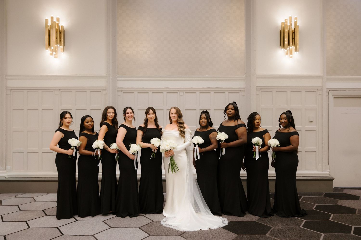Bride in white gown poses with bridesmaids in black dresses holding white bouquets in an elegant ballroom.
