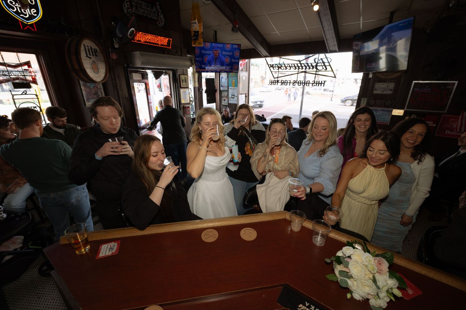 A bride in a white dress celebrates with a group of women at a bar, smiling and holding drinks together.