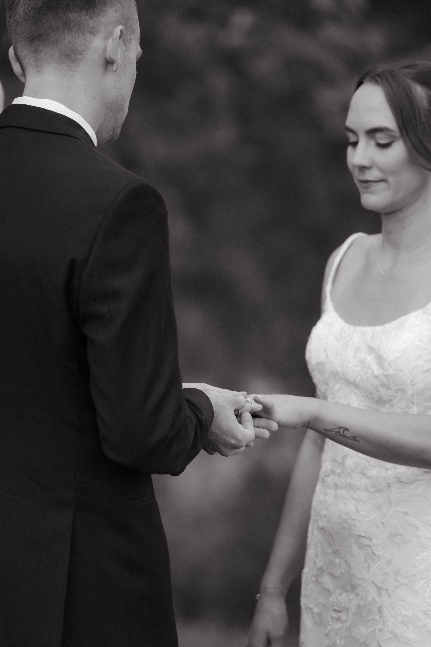 Black and white close-up of groom placing wedding ring on brides finger during outdoor ceremony.