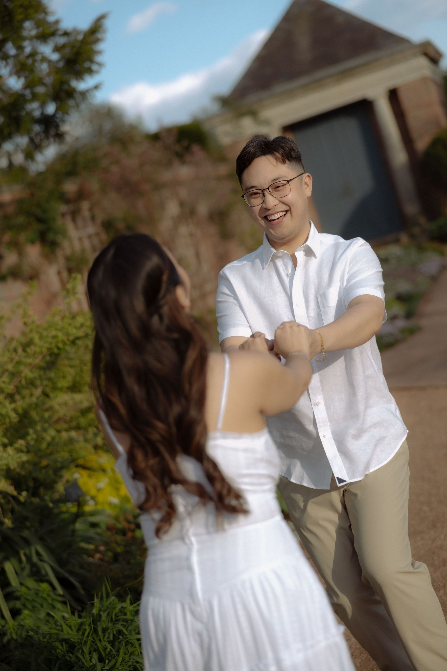 A smiling man in glasses and white shirt holds hands with a woman in a white dress outdoors.