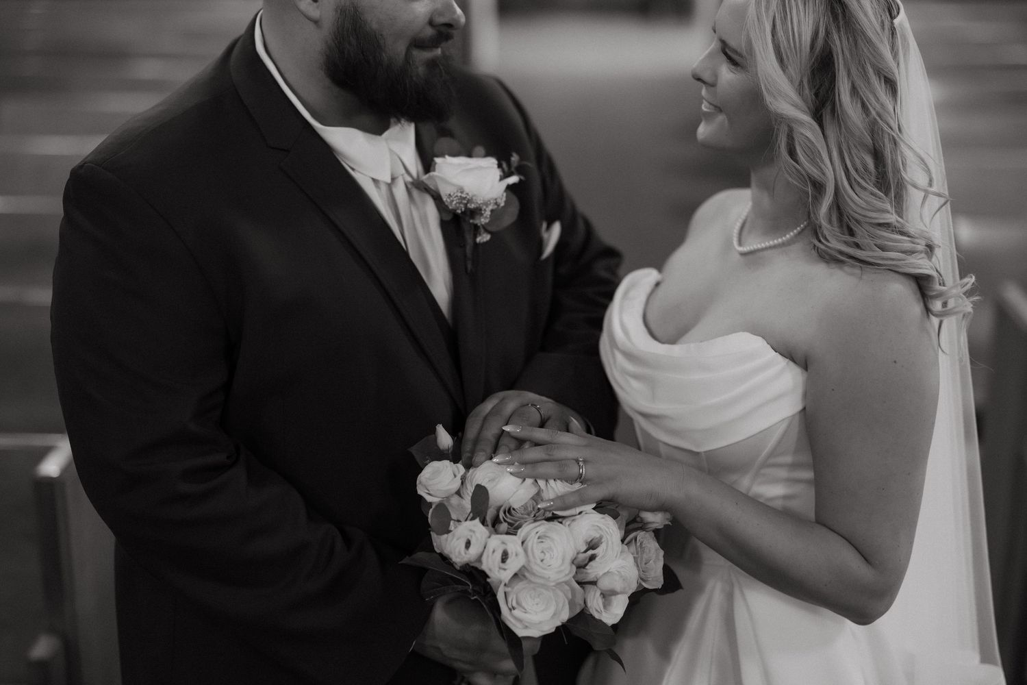 Black and white wedding photo of smiling bride holding bouquet and groom in suit exchanging rings at church ceremony.