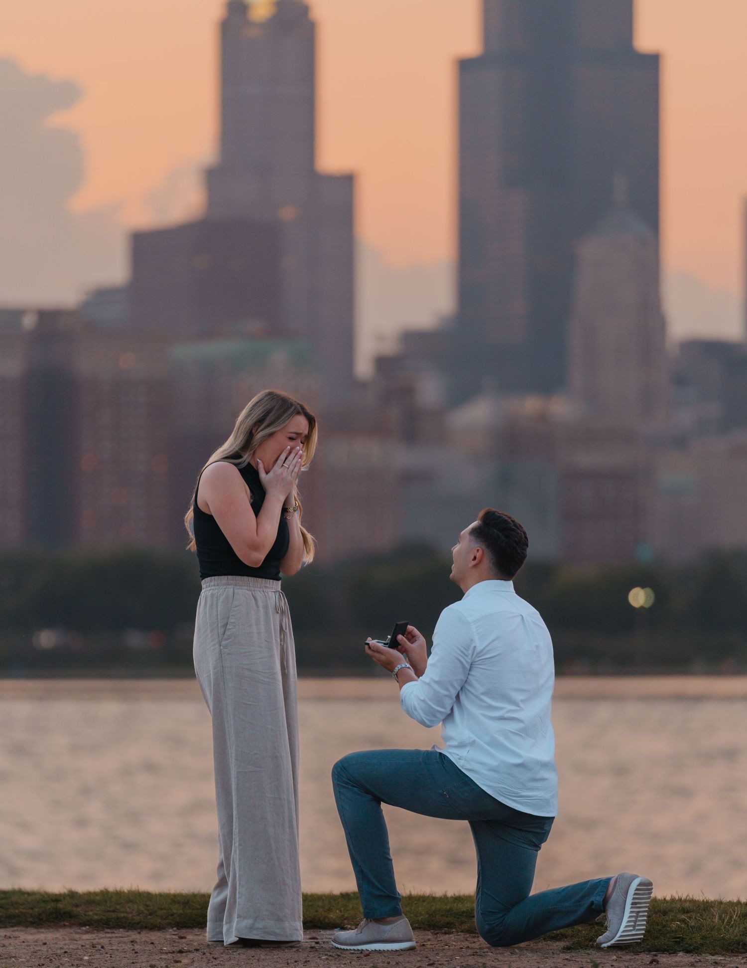 Man down on one knee proposing to his emotional girlfriend with the Chicago skyline at sunset.