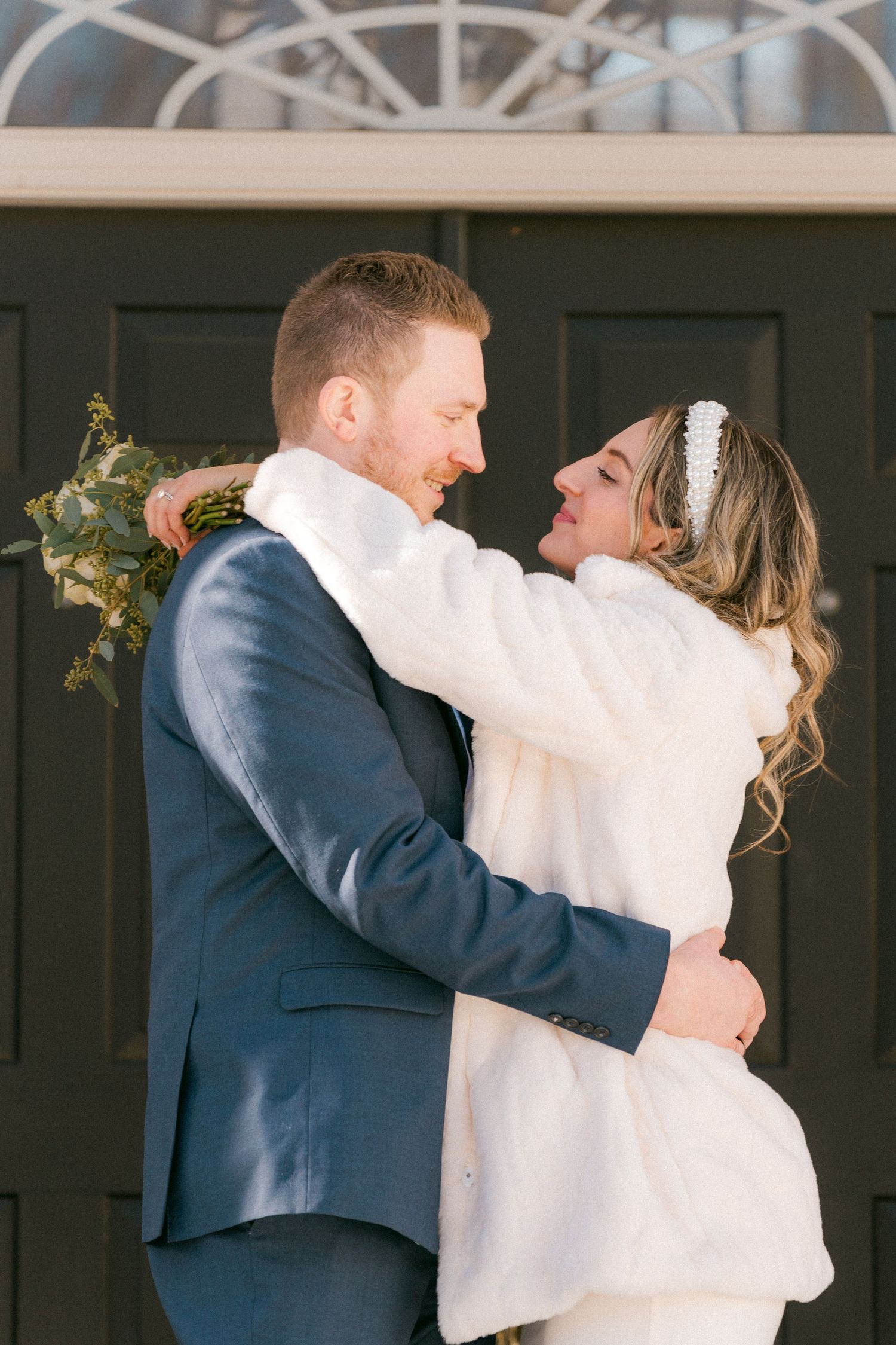 Bride in white fur coat embraces groom in navy suit outside black door in winter setting.