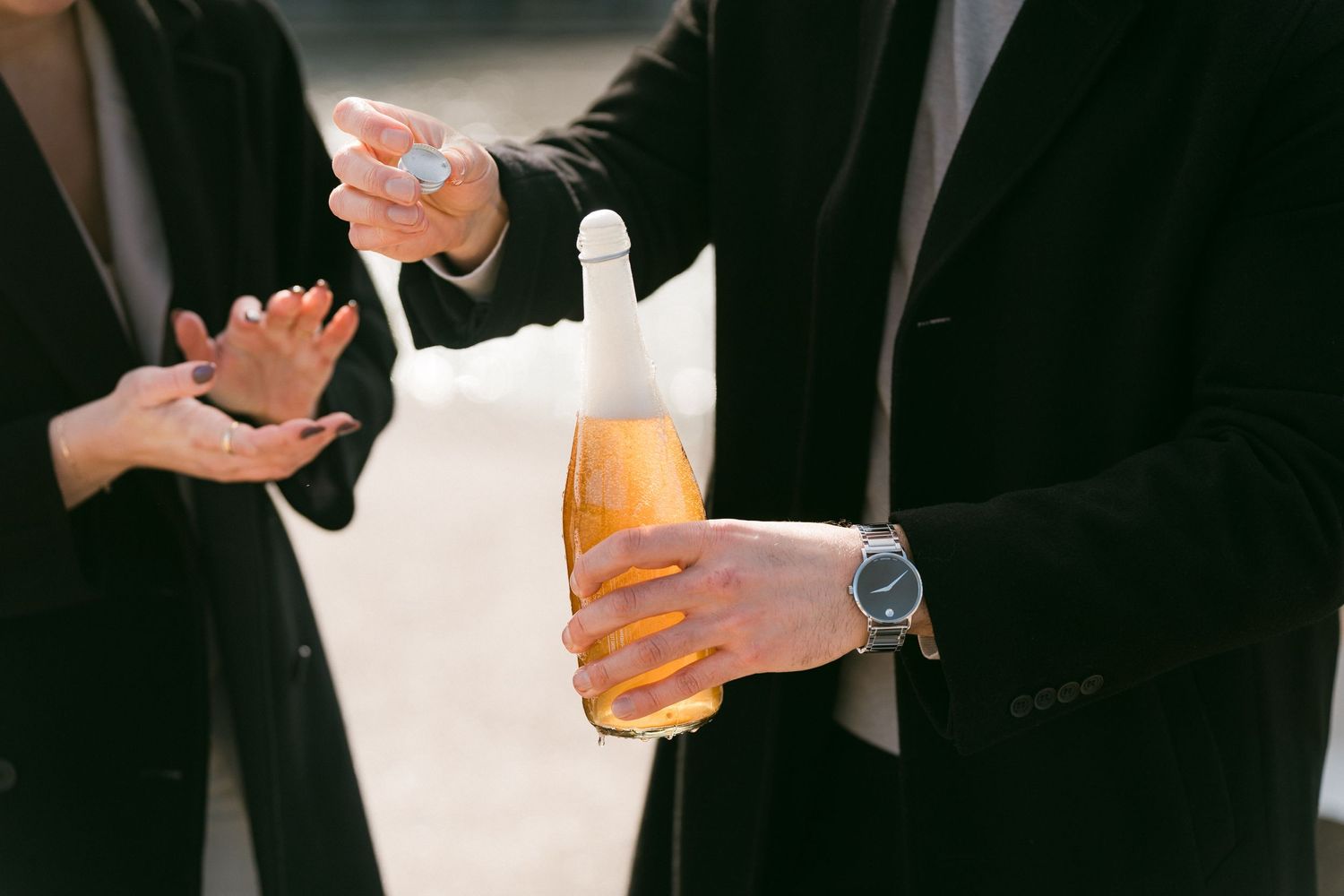 Close-up of man holding rosé champagne bottle with watch while woman gestures nearby.