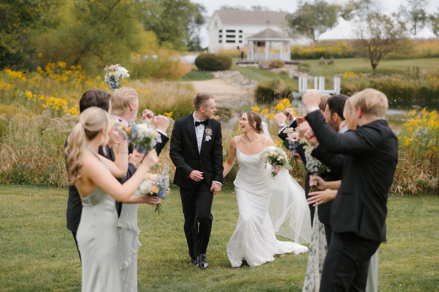 Bride and groom walk through cheering wedding party in outdoor field with wildflowers and rustic farmhouse backdrop.