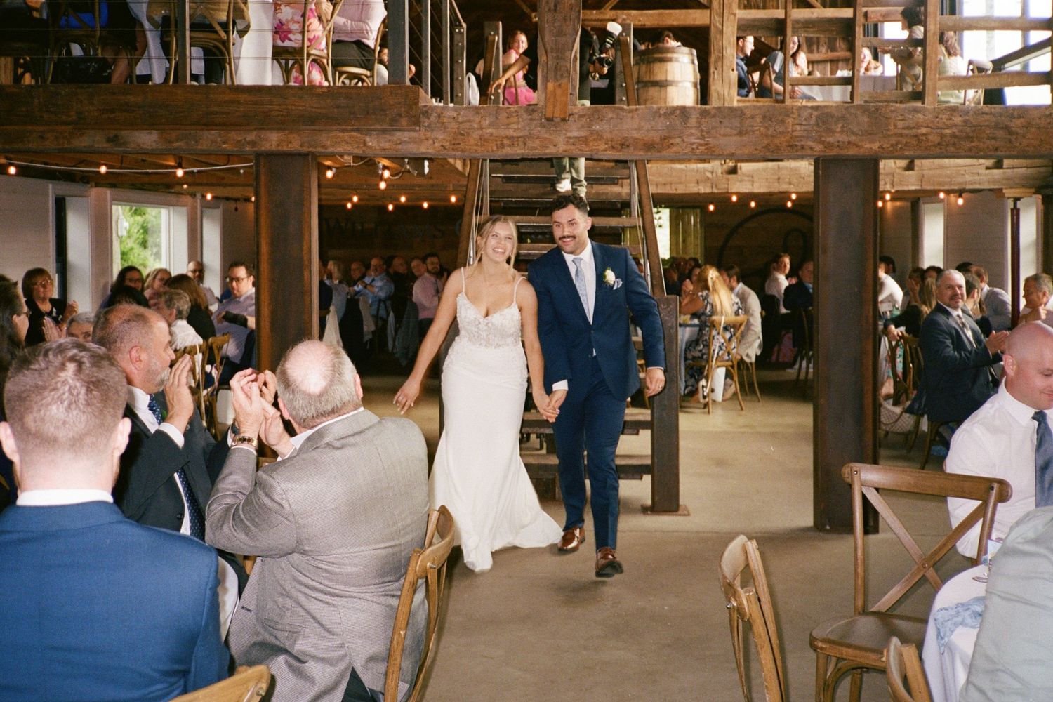 Bride and groom walk hand-in-hand down the aisle in a rustic barn wedding venue with string lights and guests.