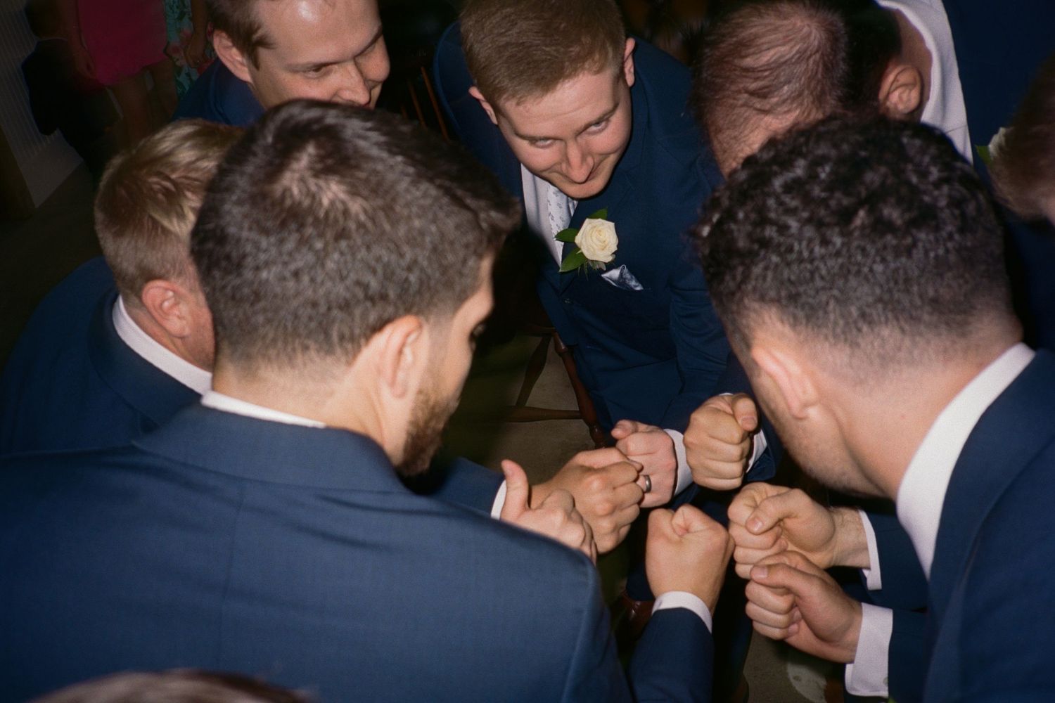 Groomsmen in navy suits forming a circle and joining hands in a celebratory huddle at a wedding reception.
