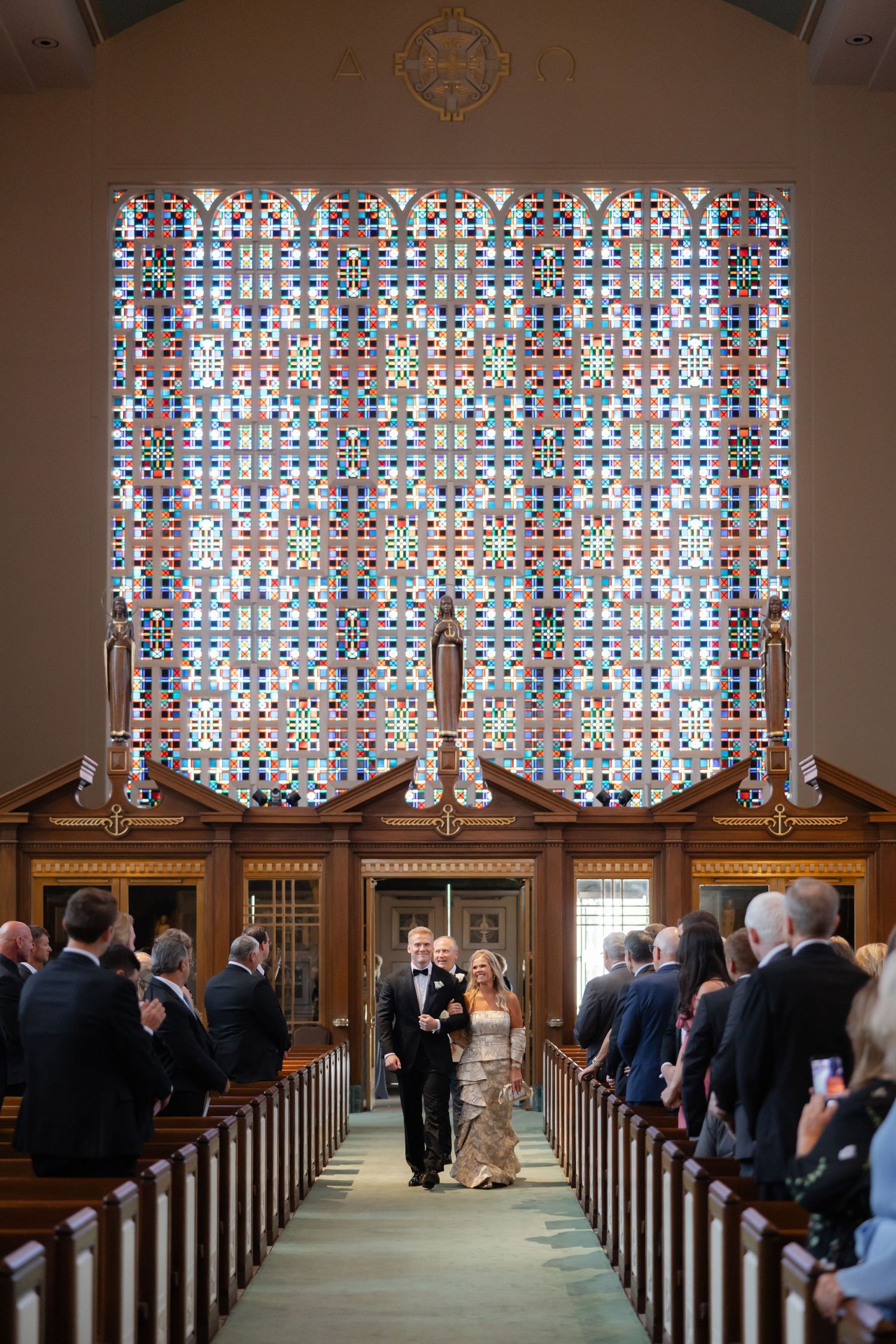 Newlyweds walk down church aisle beneath stunning colorful stained glass window as guests applaud.