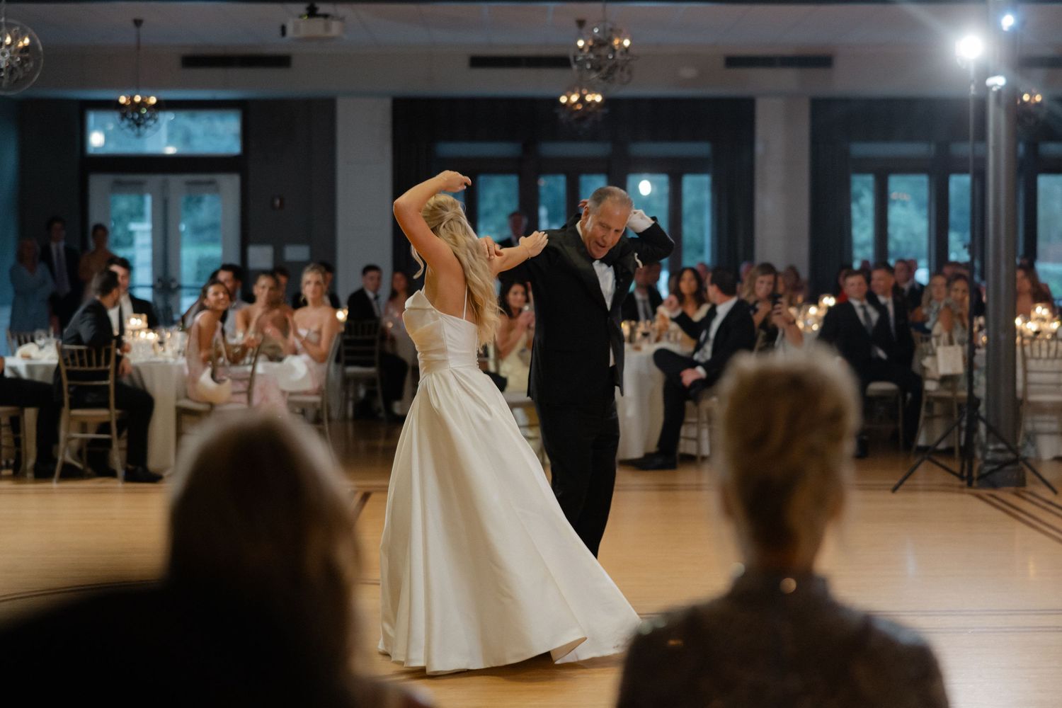 Bride and groom share first dance in elegant ballroom while wedding guests watch and applaud.