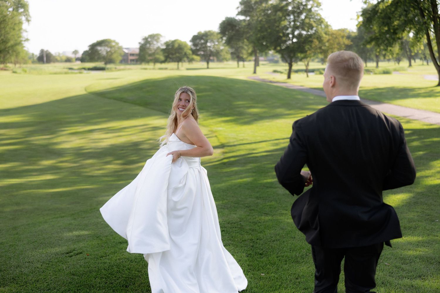 Smiling bride in white gown twirls dress while groom watches on scenic golf course.