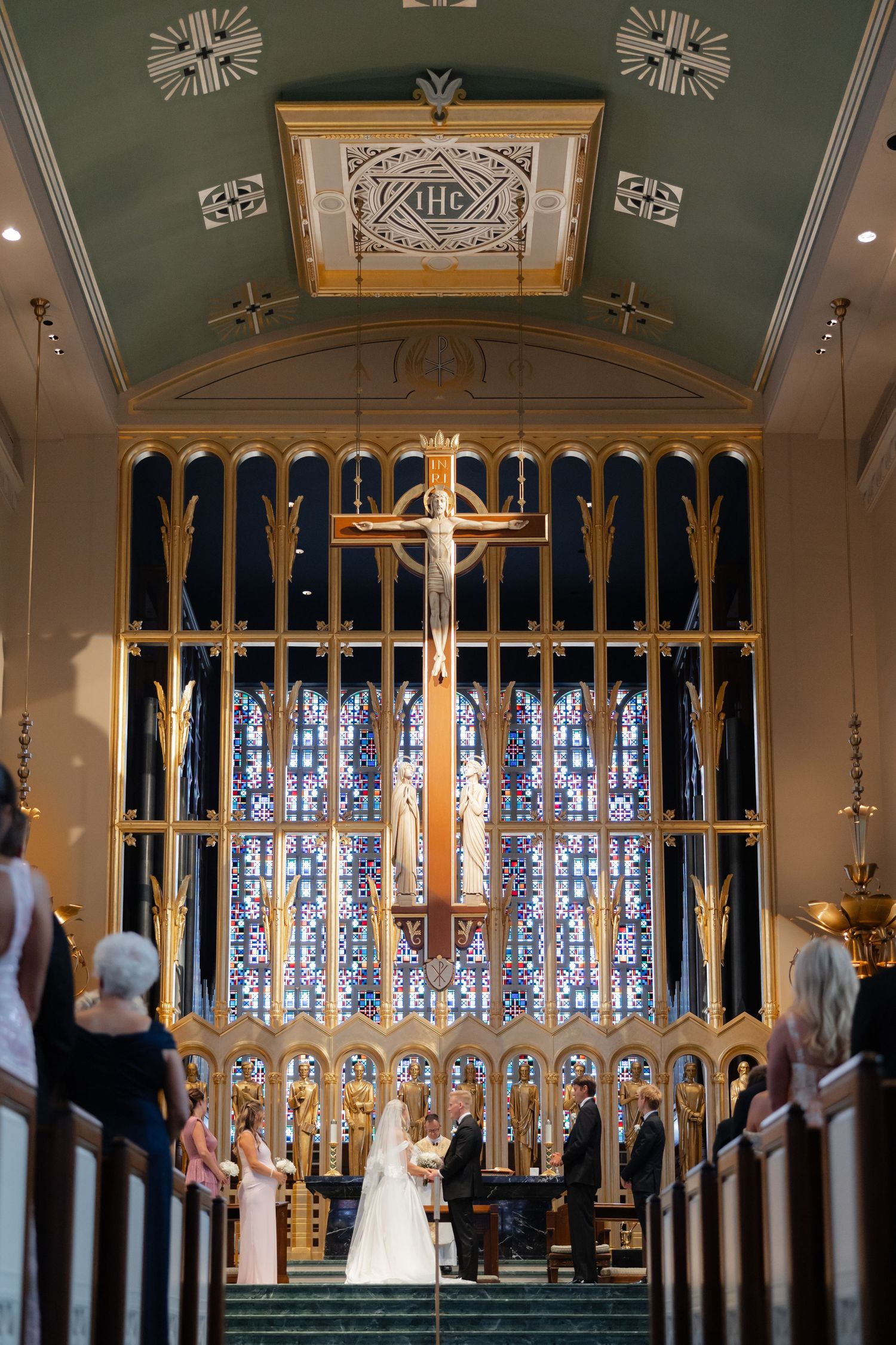 Bride and groom exchange vows at ornate church altar with golden crucifix and stunning stained glass windows.