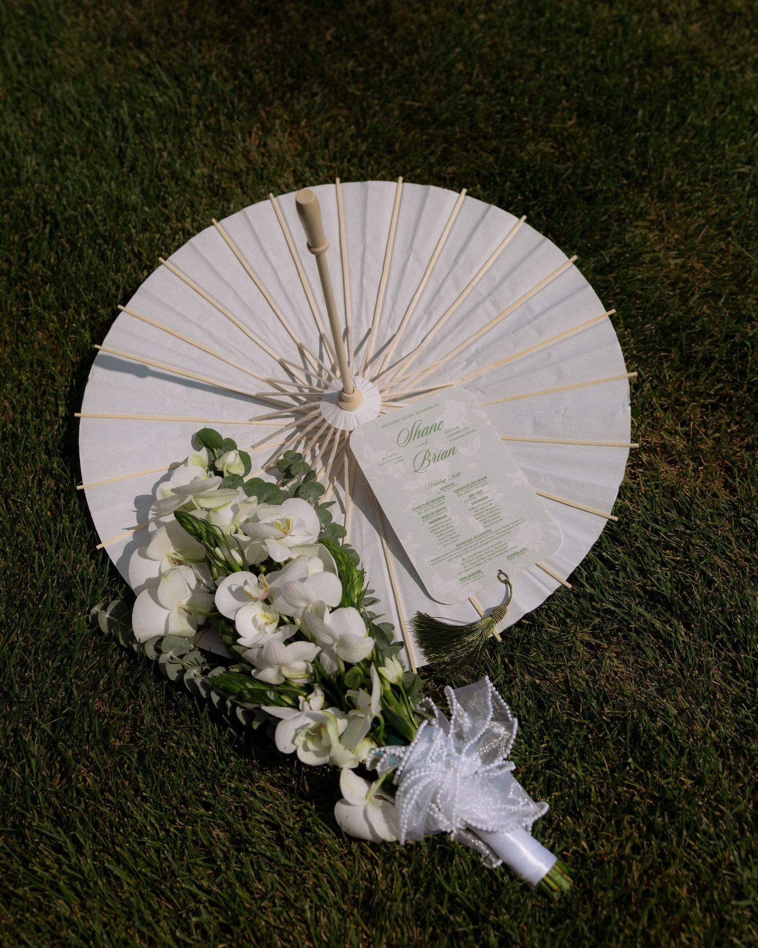 White parasol umbrella with wedding program and white floral bouquet with ribbon on green grass.