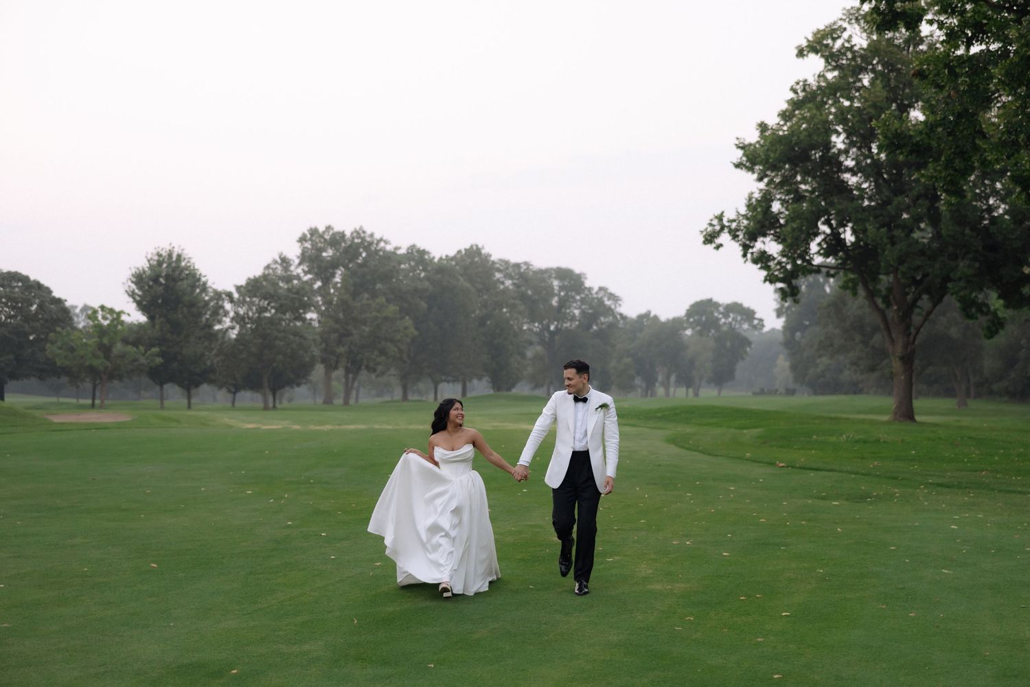 Bride and groom holding hands walking across lush green golf course at dusk.