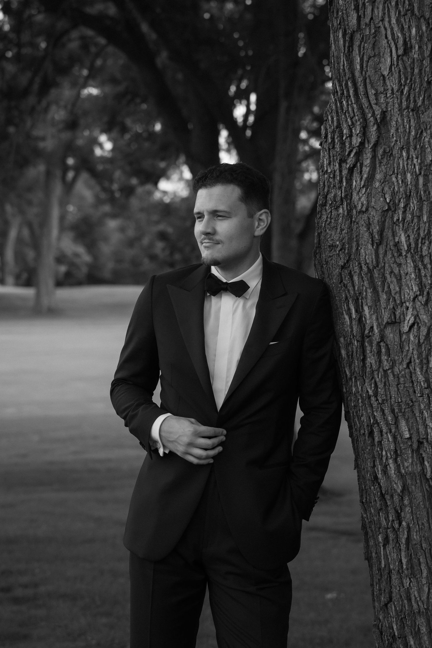 Black and white photo of a man in a tuxedo with bow tie leaning against a tree in a park.