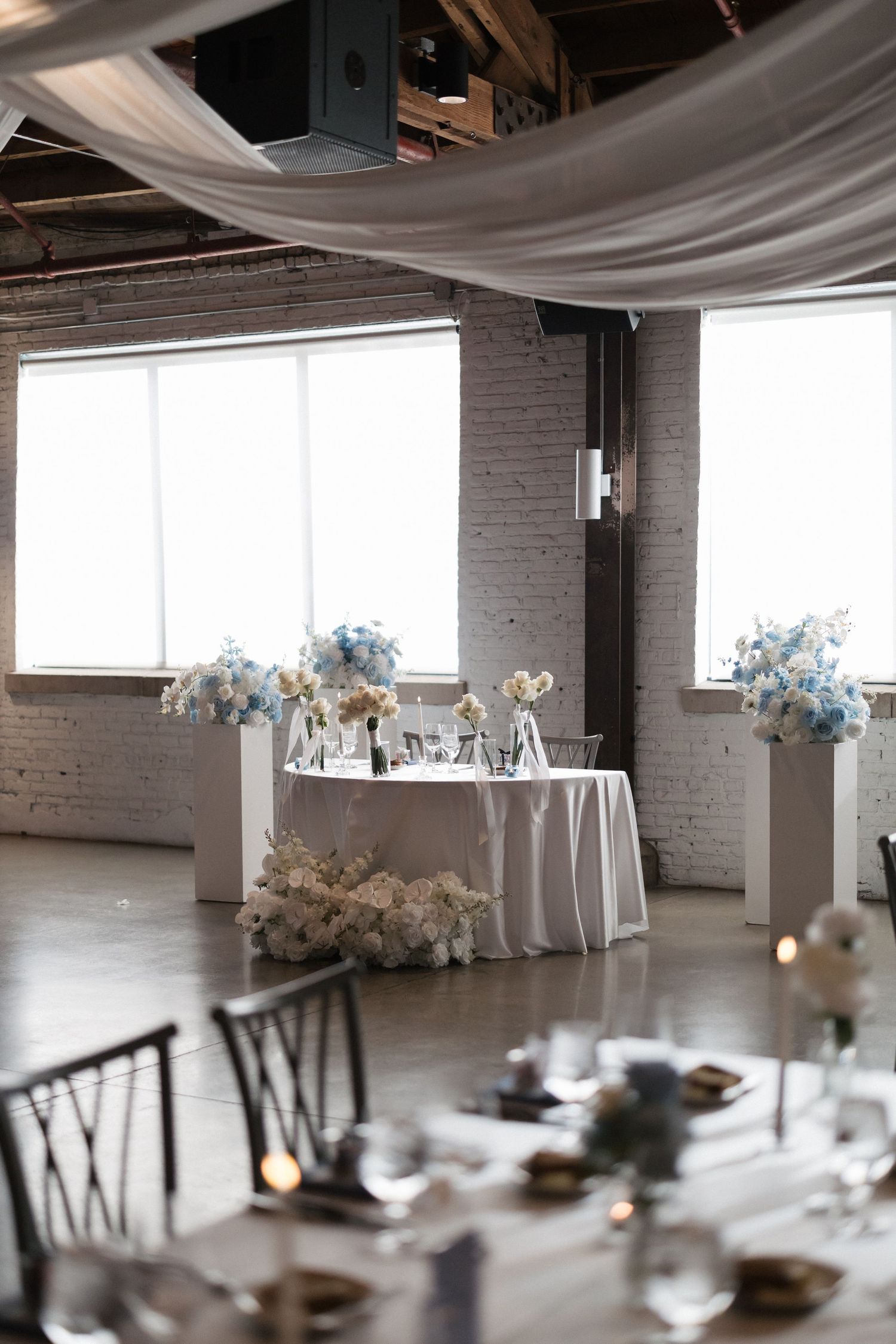 Elegant wedding reception venue with white-draped sweetheart table, blue floral arrangements, and industrial brick walls.