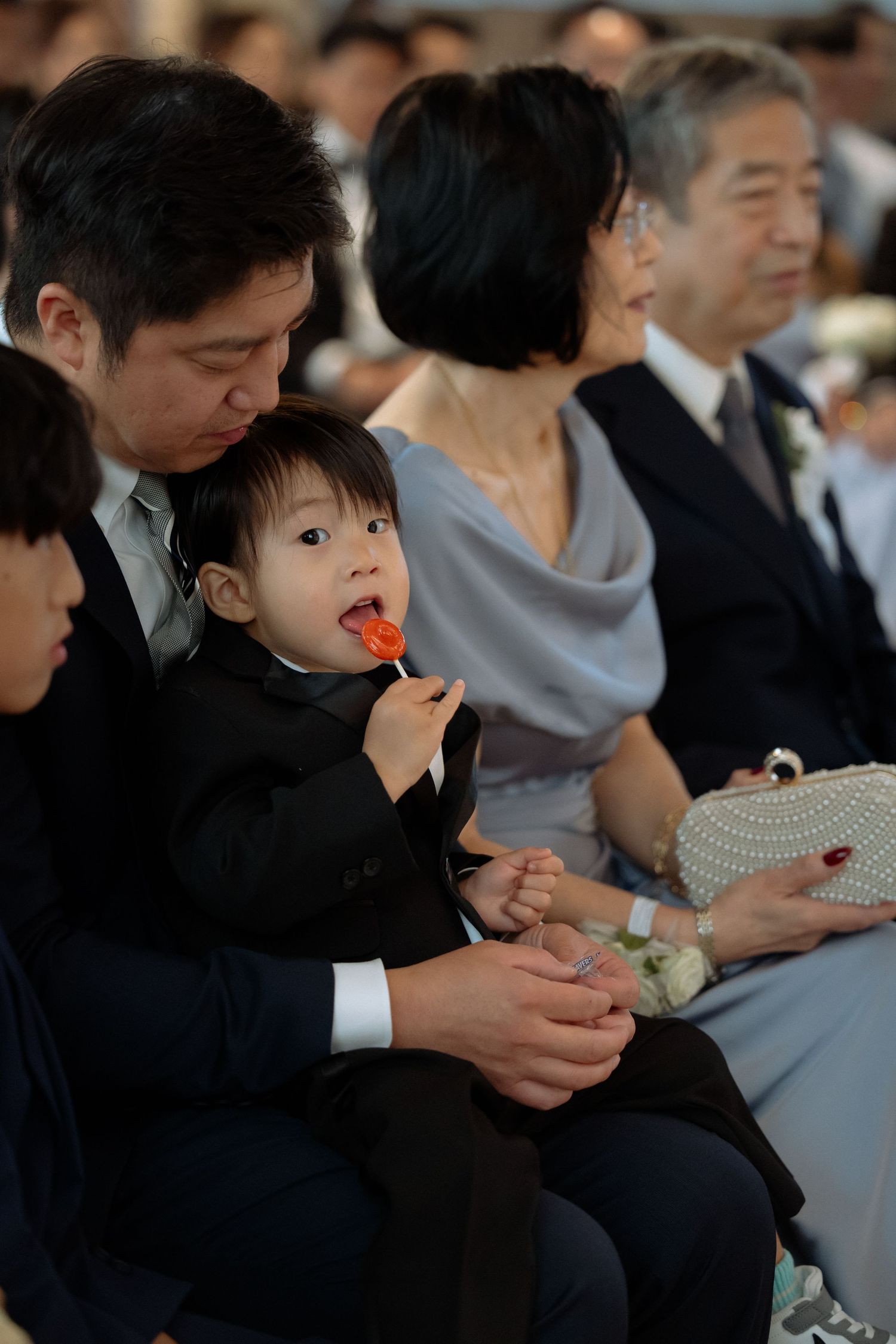 Asian family at formal event, father holding toddler in suit eating lollipop, elderly couple seated behind them.