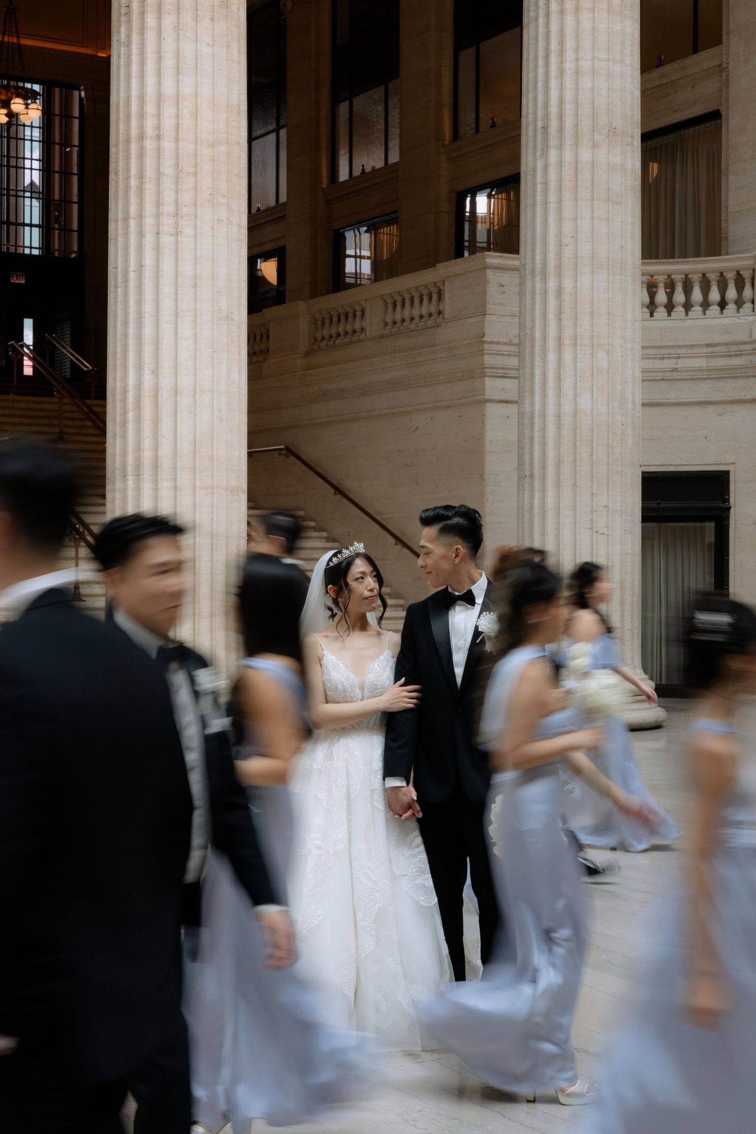 Bride and groom share an intimate moment surrounded by blurred wedding party in a grand columned hall.