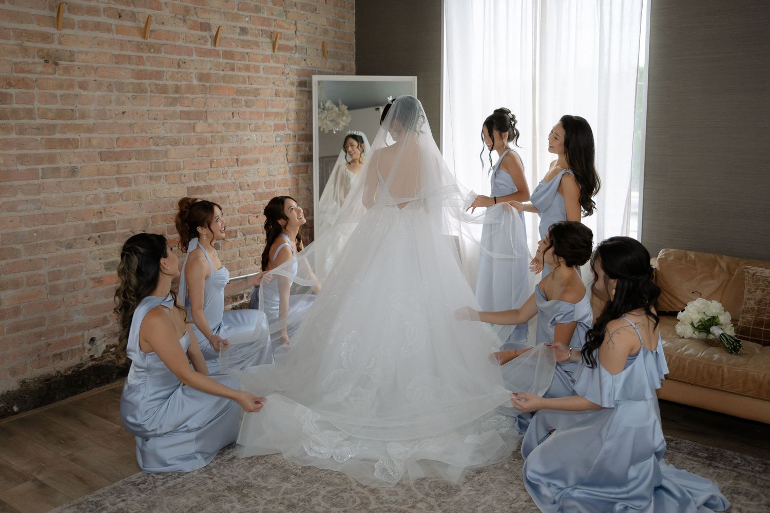 Bride in white ball gown and veil surrounded by bridesmaids in blue dresses arranging her train in a brick-walled room.