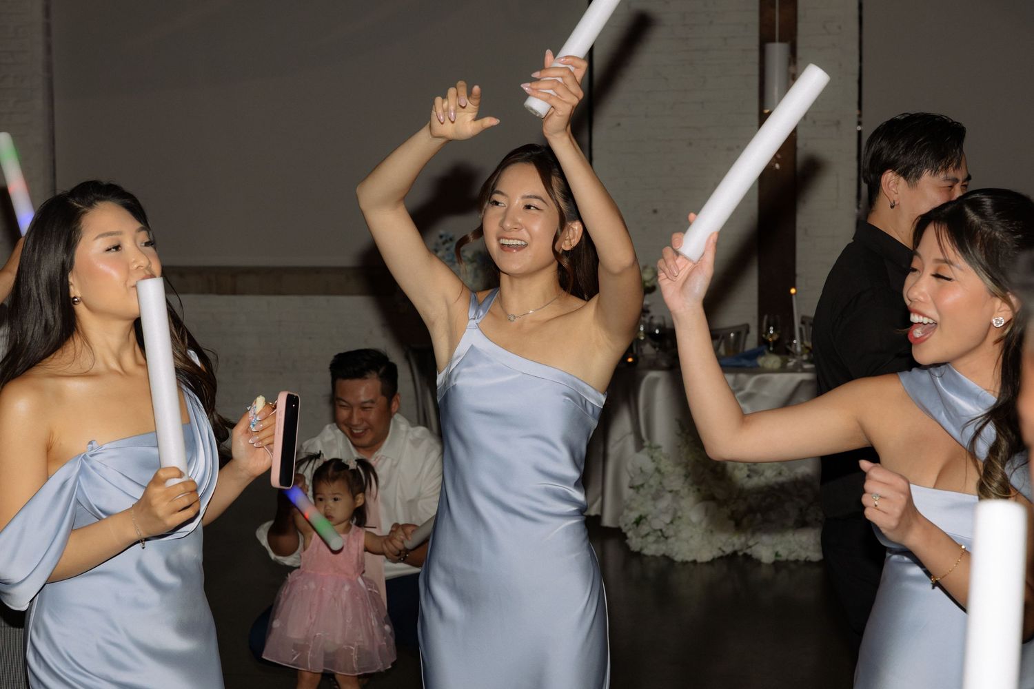 Bridesmaids in blue dresses dancing and waving glow sticks joyfully at wedding reception party.