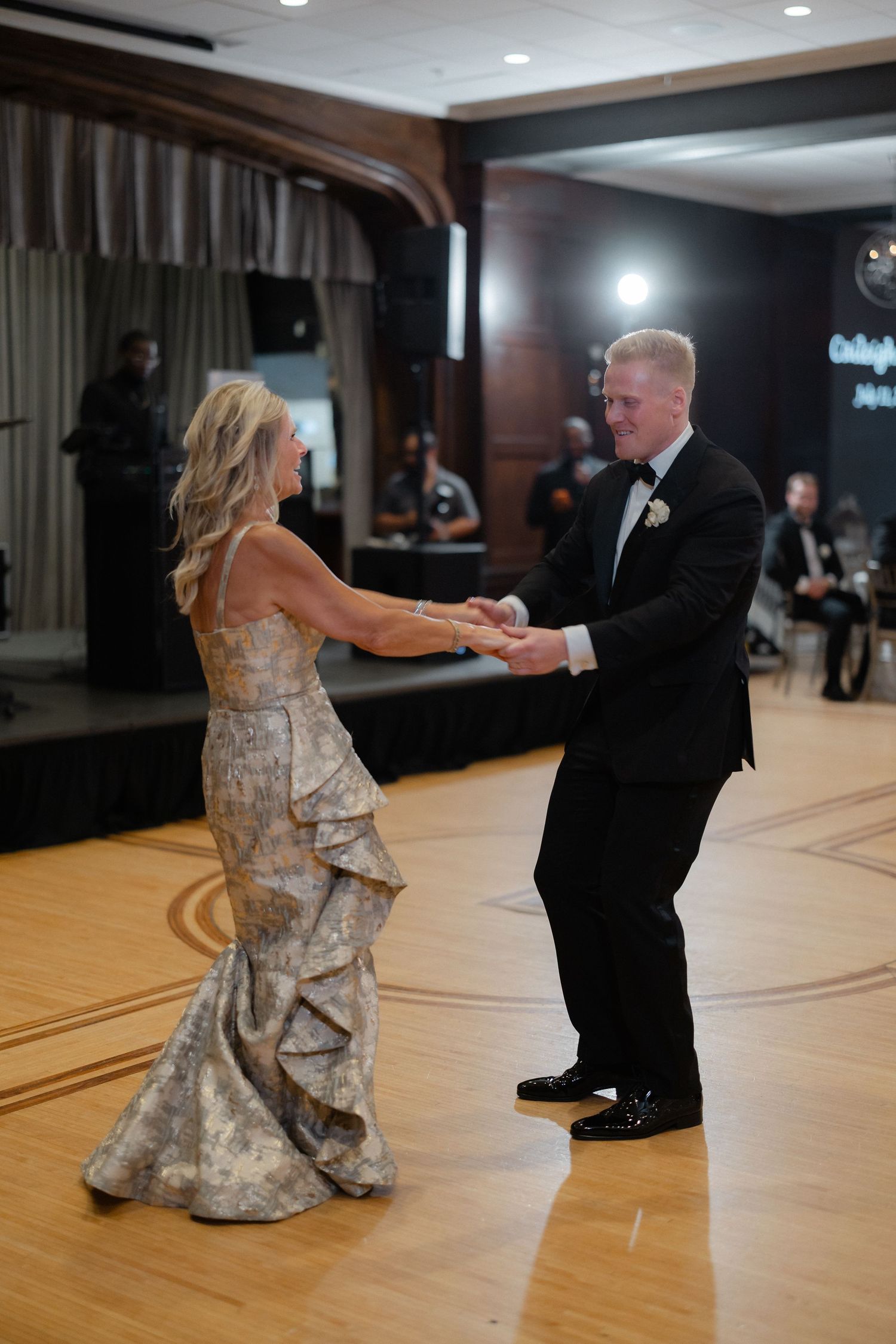 Elegant mother-son wedding dance, woman in gold gown and groom in black tuxedo on ballroom floor.