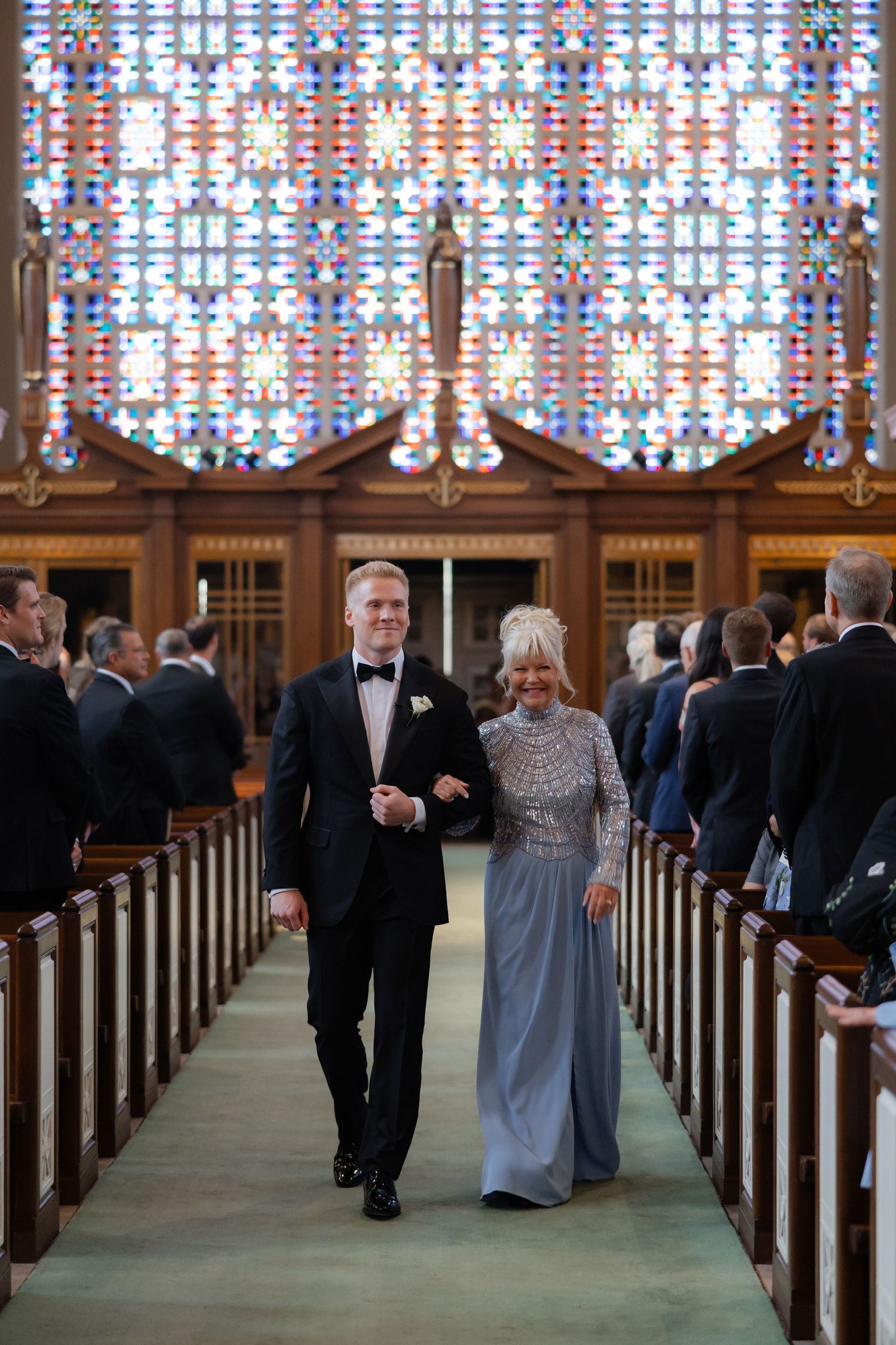Groom escorts his mother down the church aisle before a stunning stained glass window at wedding ceremony.