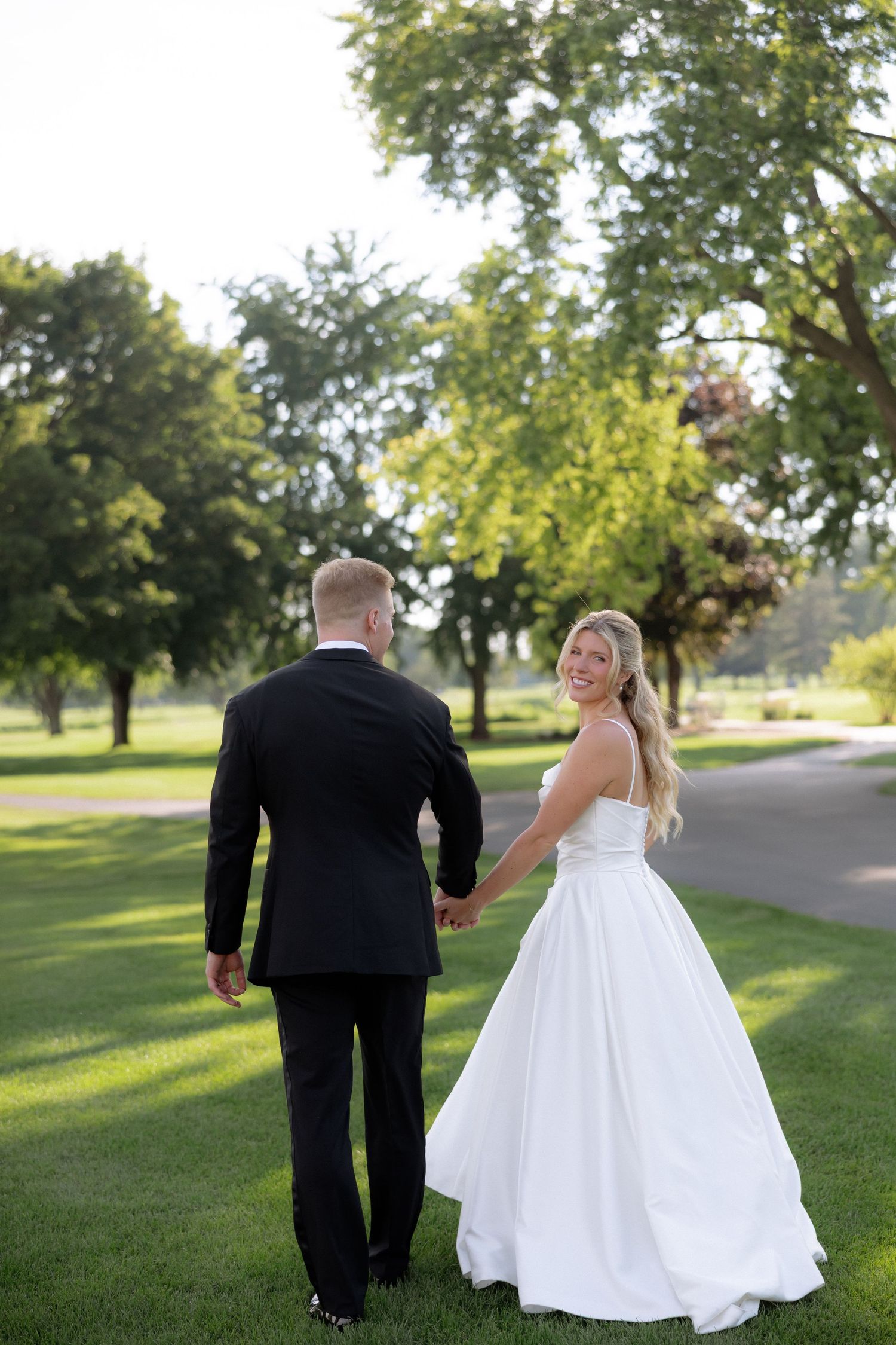 Bride in white ball gown looks back smiling while holding hands with groom walking through green park.