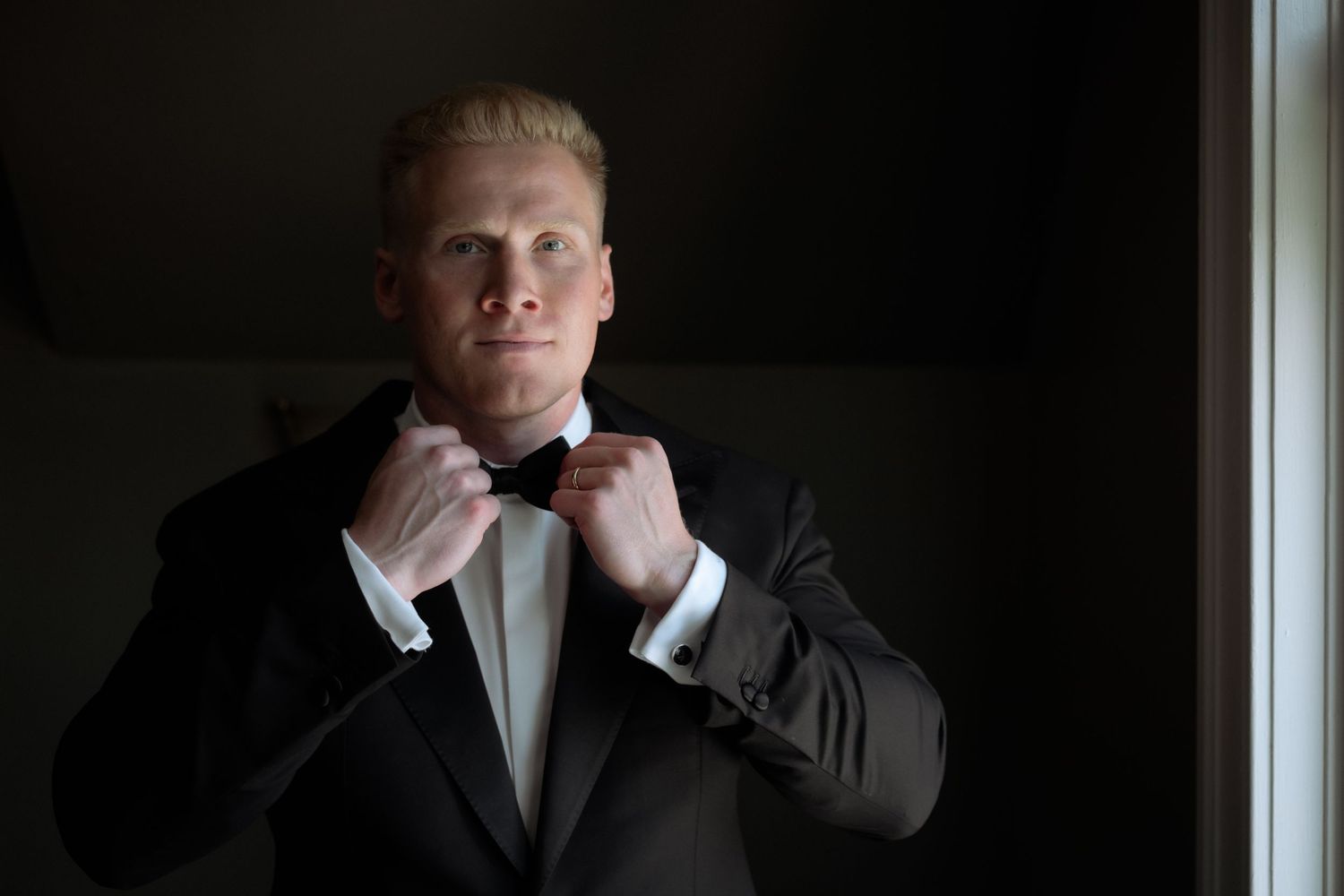 A blond groom in a black tuxedo adjusts his bow tie while standing near a window in soft natural light.