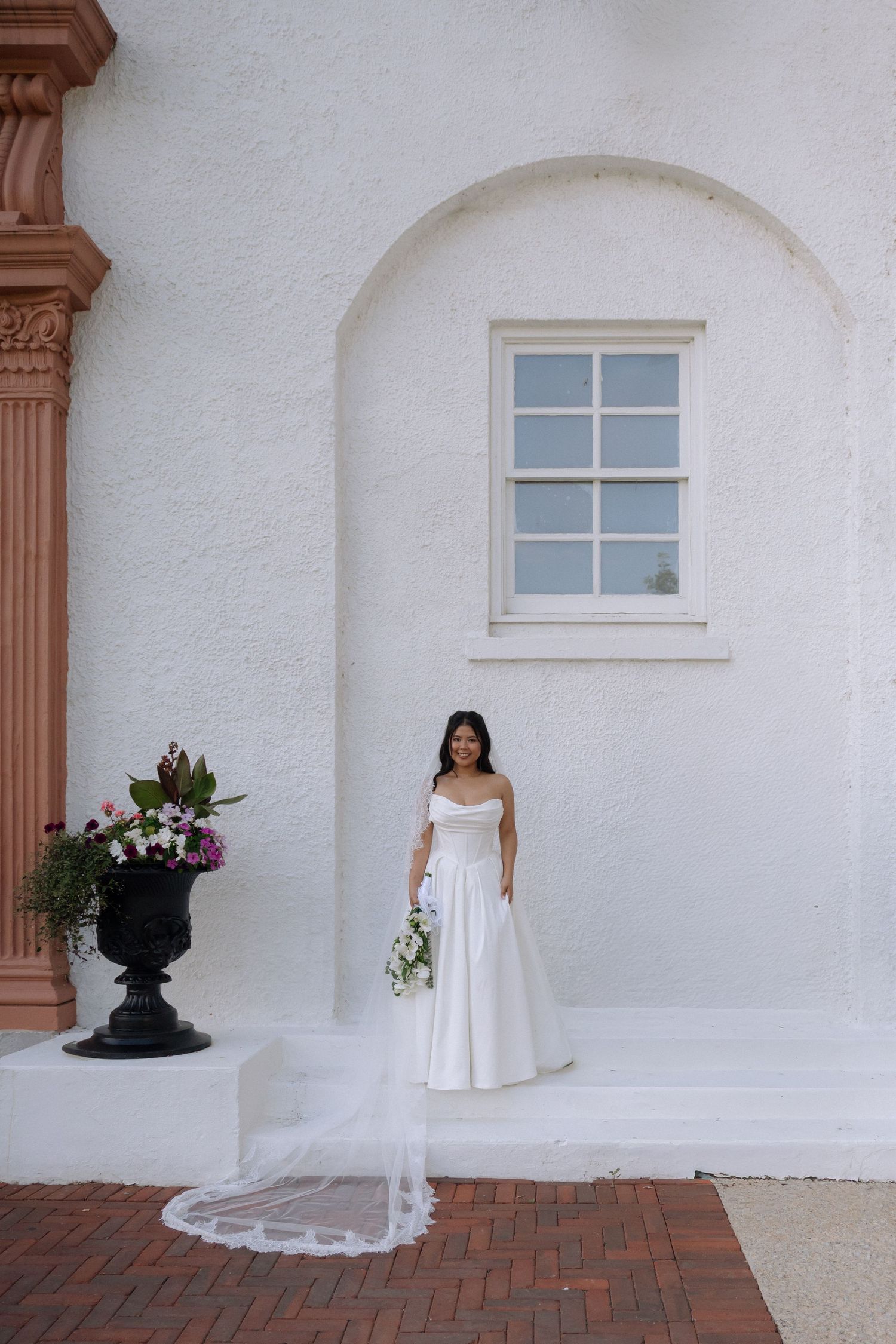 Bride in strapless white gown with cathedral veil holds bouquet outside white stucco building with arched window.