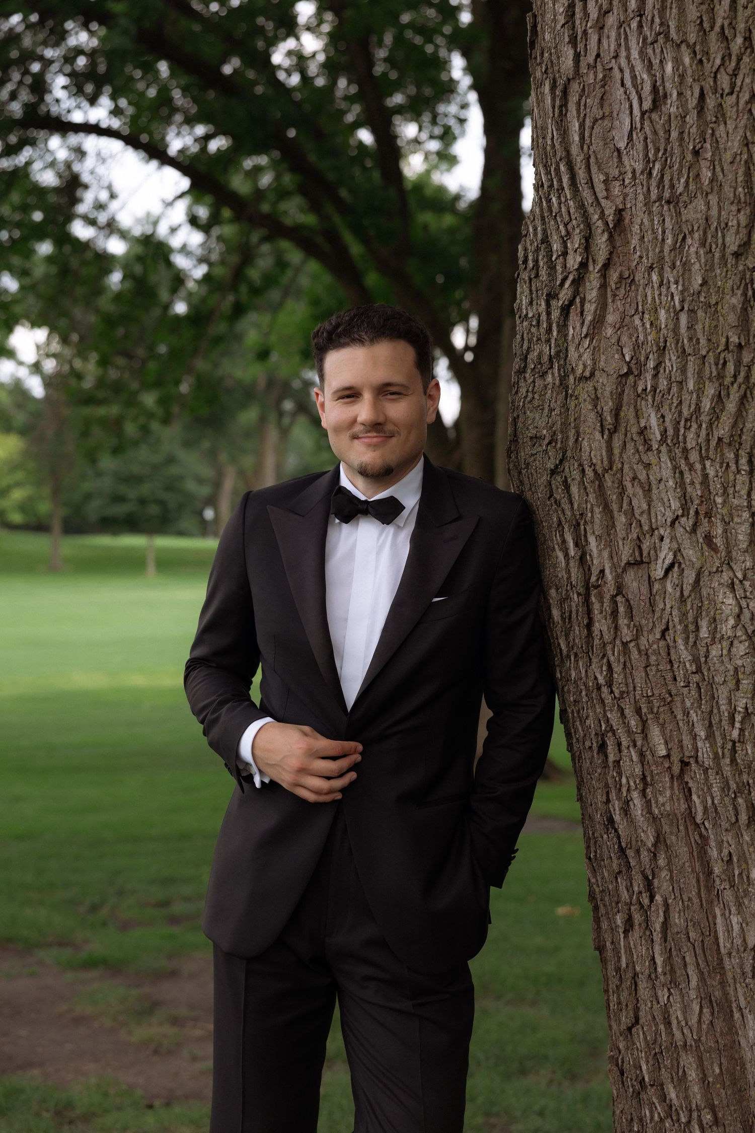 Handsome groom in black tuxedo and bow tie smiling while leaning against a tree outdoors.