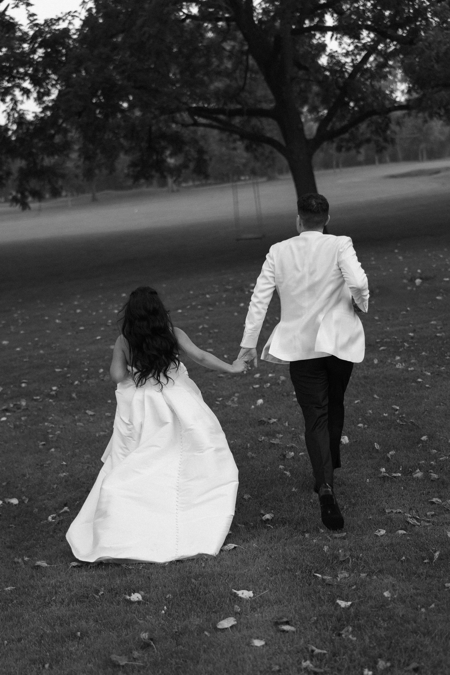 Bride and groom hold hands walking across park lawn, black and white wedding portrait from behind.