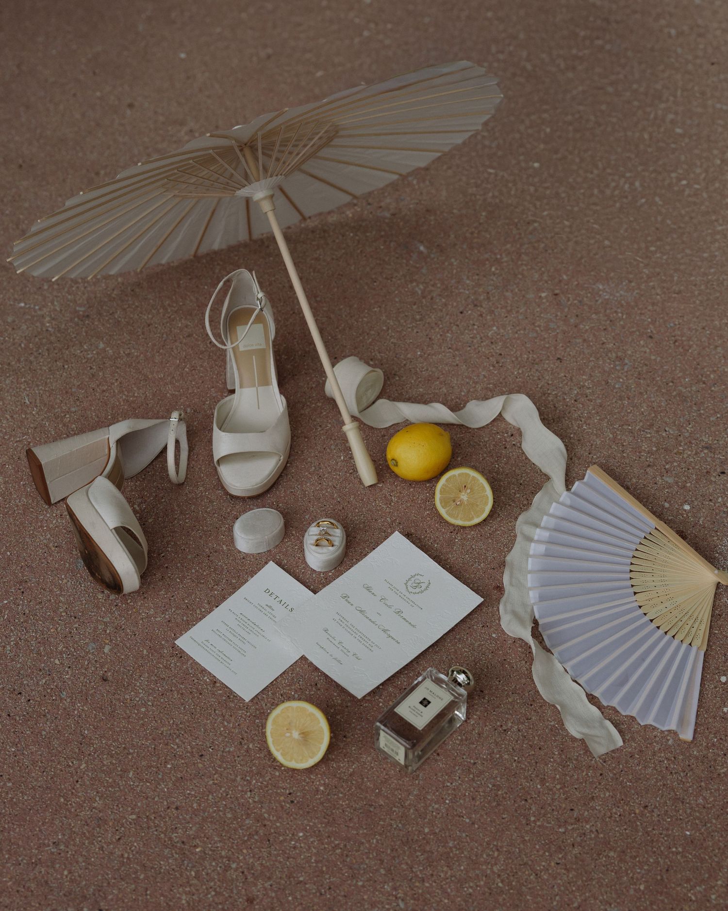 Elegant wedding flat lay with white heels, parasol, paper fan, lemons, stationery, and Jo Malone perfume on sandy surface.