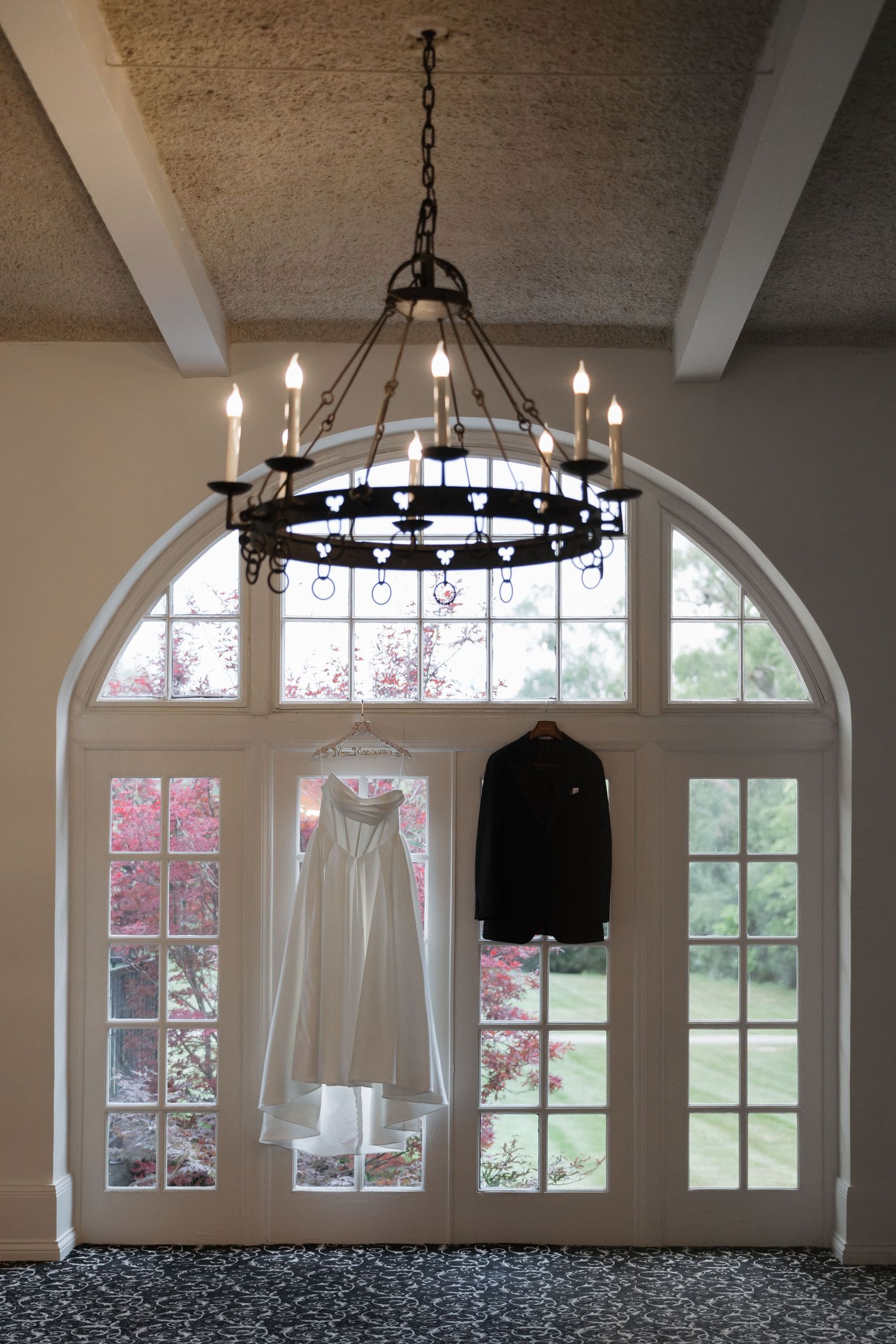 Wedding dress and groom suit hanging from elegant black chandelier in arched window foyer of wedding venue.