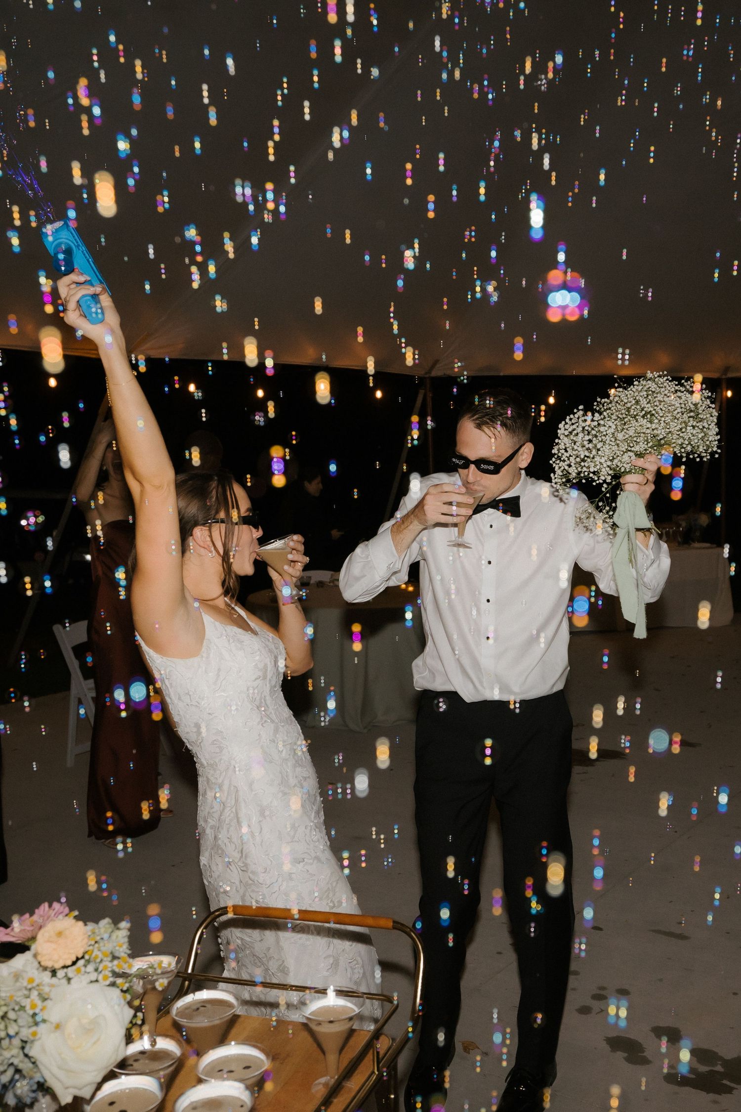 Bride and groom celebrating at wedding reception with colorful bokeh lights, champagne toast, and bridal bouquet.