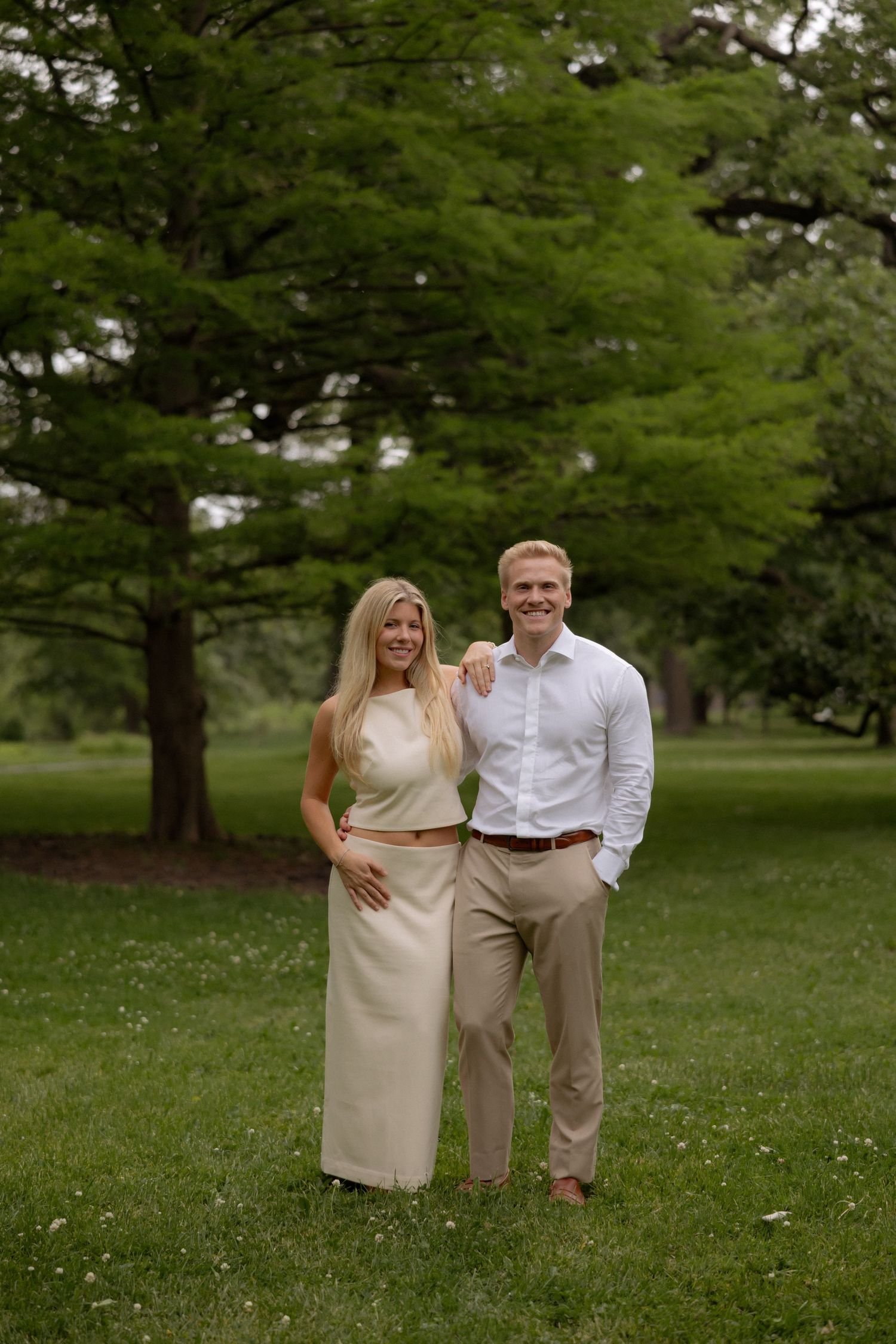 Smiling couple standing together on green grass in a park, woman in cream outfit, man in white shirt.