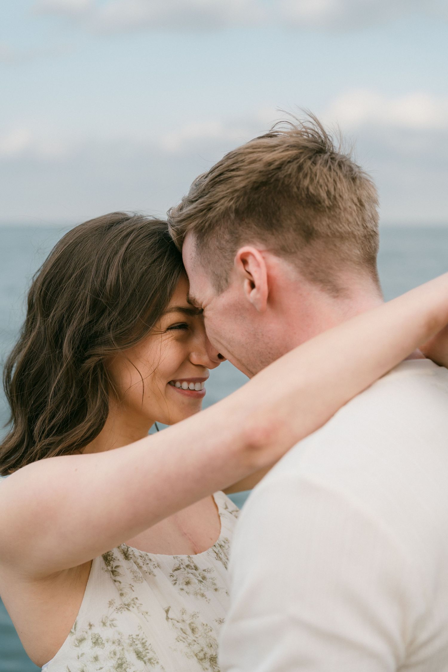 Smiling woman embracing man closely near ocean with cloudy blue sky backdrop.