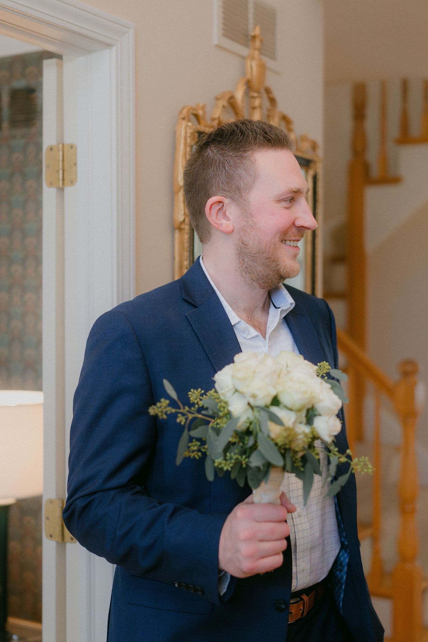 Smiling groom in navy suit holds white rose bouquet while standing in elegant home entryway.