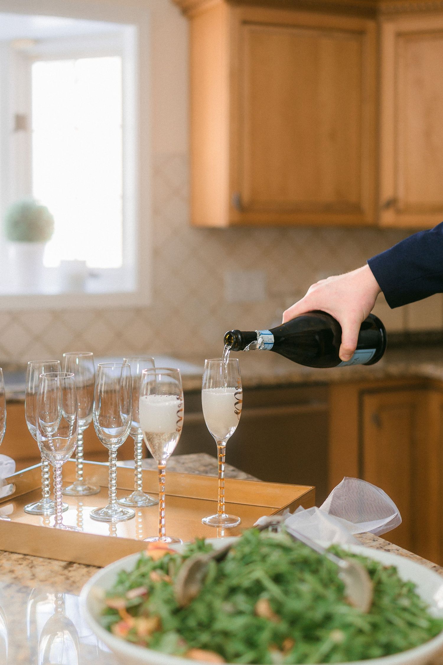 Hand pouring champagne into flute glasses on a gold tray in a kitchen beside a fresh arugula salad.