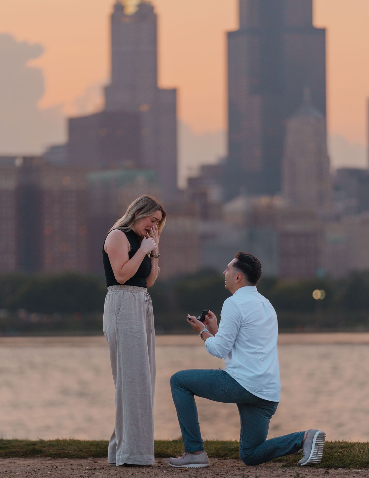 Man proposing on one knee to his tearful girlfriend against the glowing Chicago skyline at dusk.