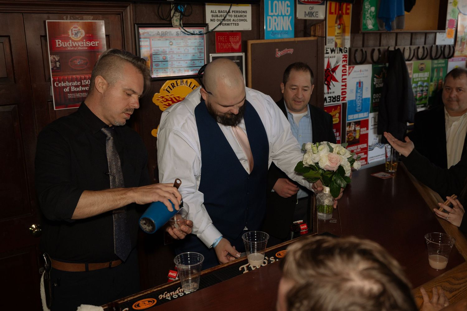 Men in formal attire gather at a bar, one pouring a drink, with white roses and beer taps visible in the background.