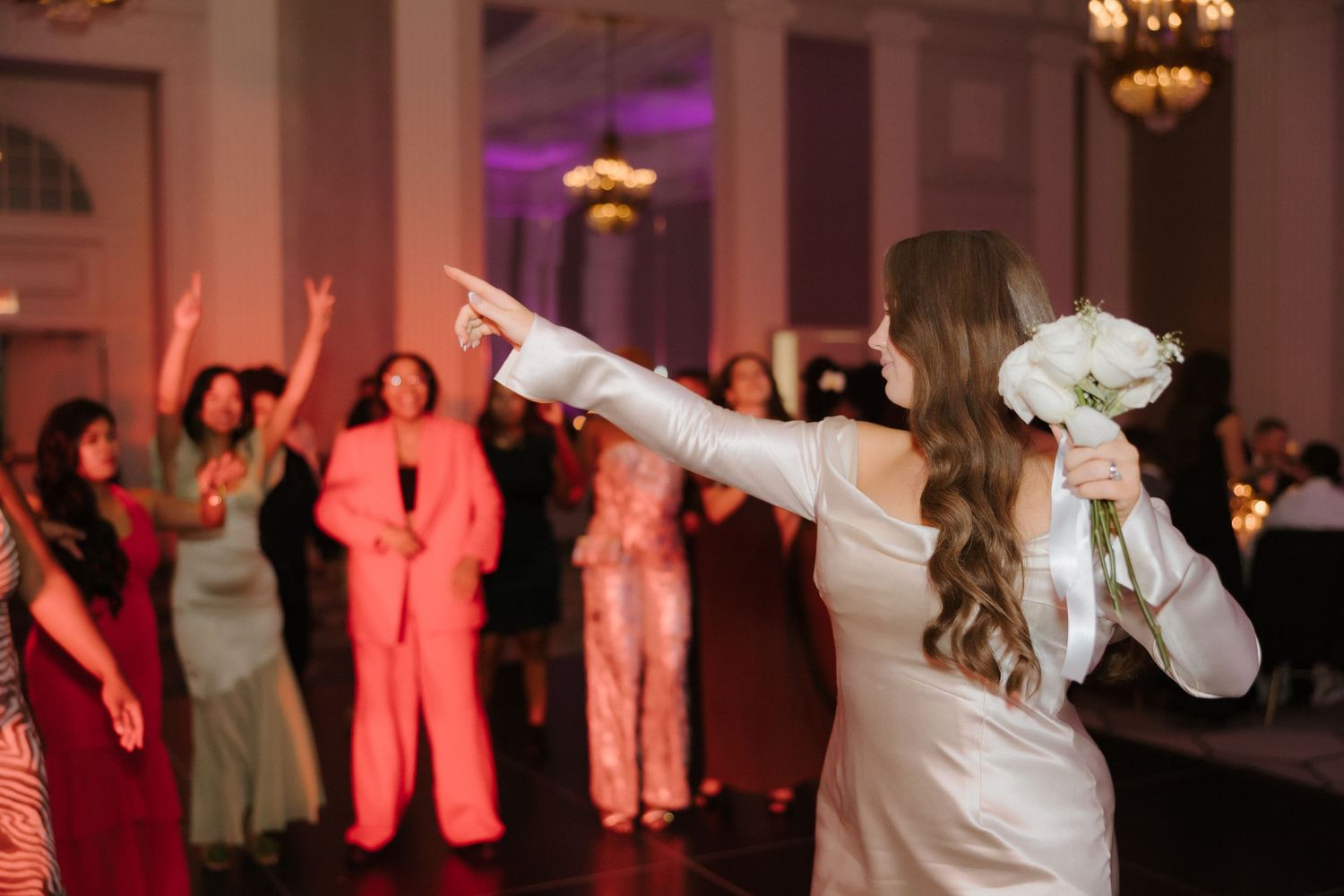 Bride in white gown holds bouquet, pointing at cheering guests on dance floor at elegant wedding reception.
