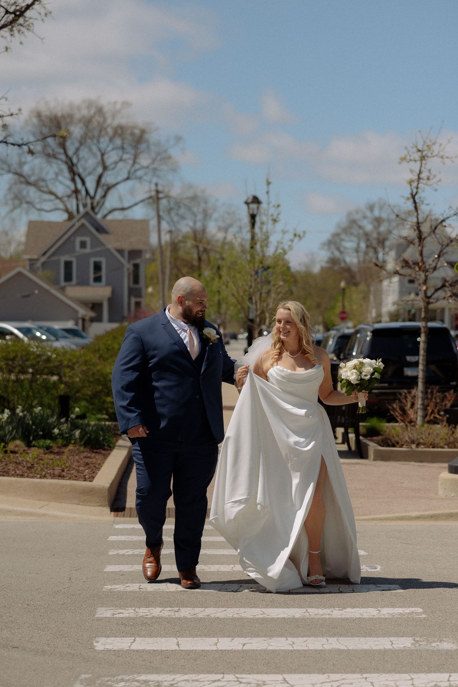Bride and groom crossing a street in a charming town, bride in white gown holding bouquet, groom in navy suit.