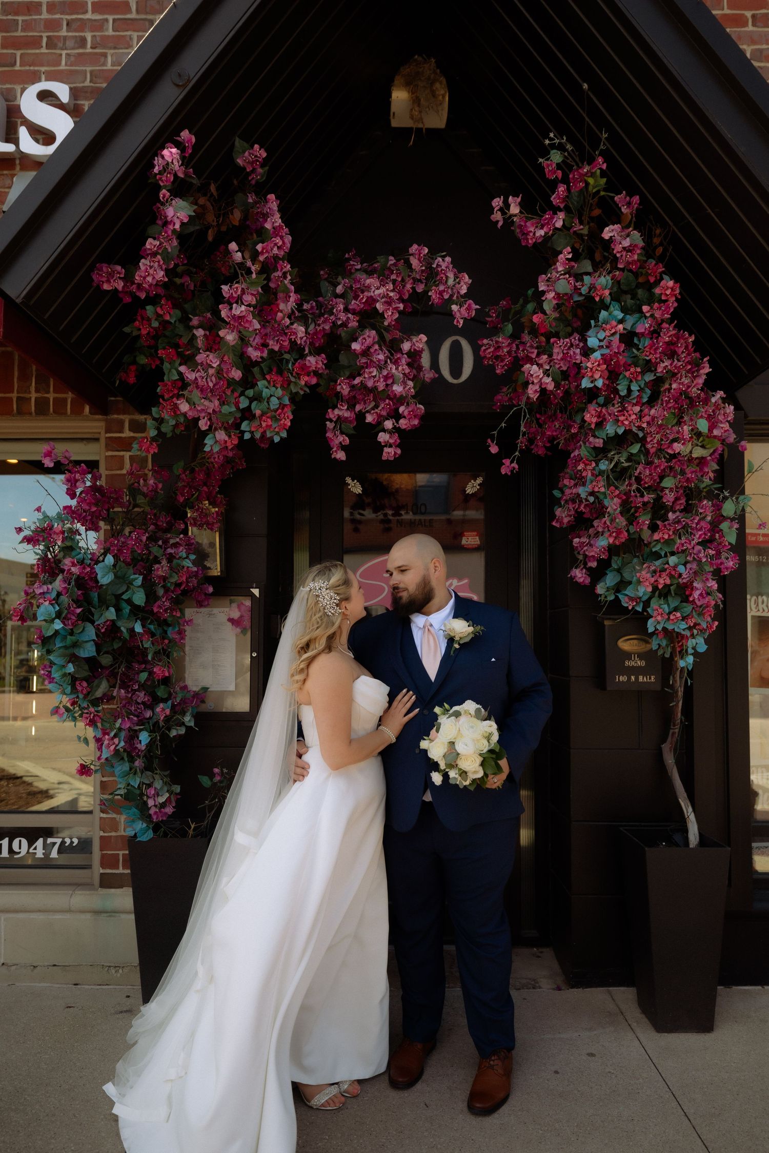 Bride and groom share a romantic moment under a floral arch adorned with pink blooms at a charming brick building entrance.