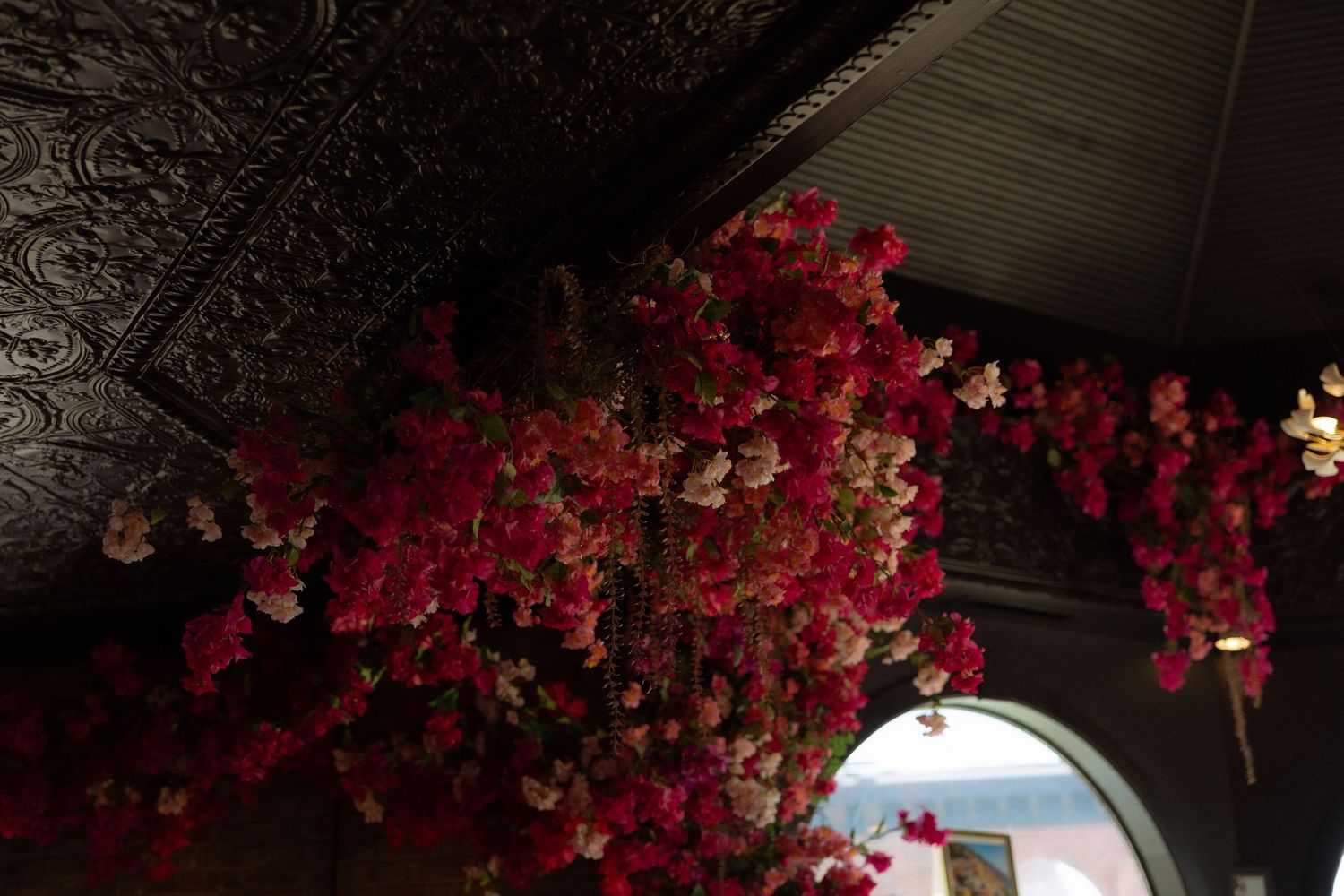 Lush pink and red floral arrangement cascading from a dark ornate tin ceiling in a dimly lit interior space.