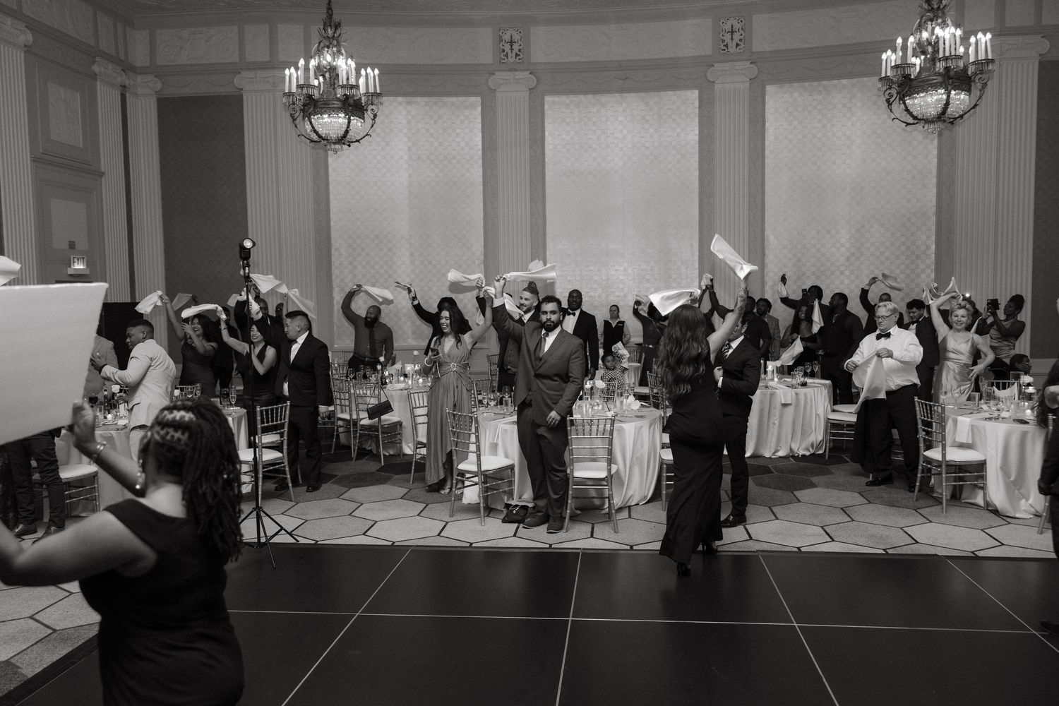 Black and white wedding reception photo showing guests celebrating with raised hands in an elegant ballroom.