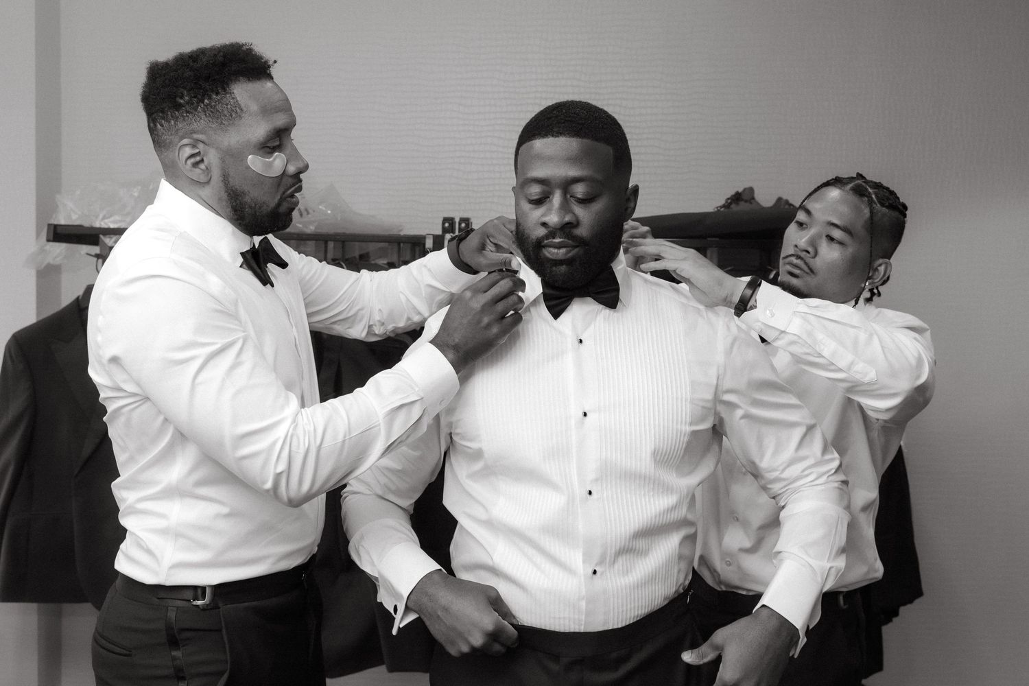 Two groomsmen help a groom adjust his bow tie and collar in a black and white wedding preparation photo.