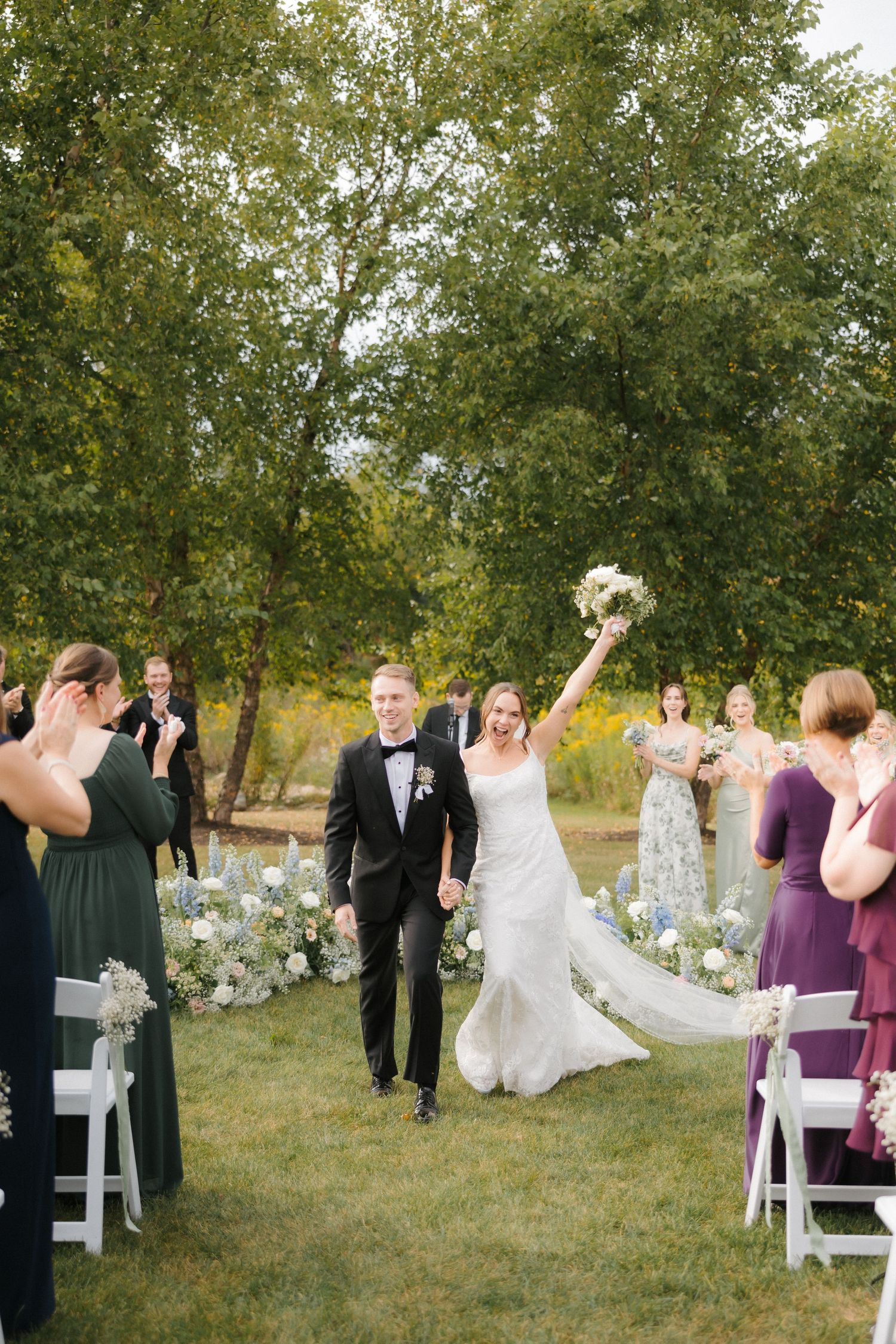 Bride raises bouquet triumphantly while walking recessional with groom past cheering guests outdoors.