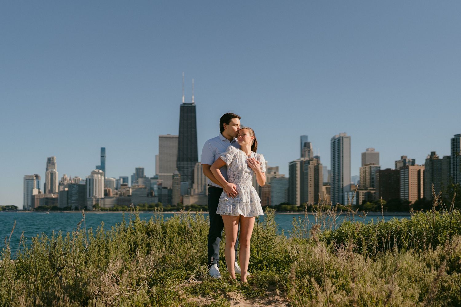 Engaged couple embracing near Lake Michigan with iconic Chicago skyline and John Hancock Tower in background.